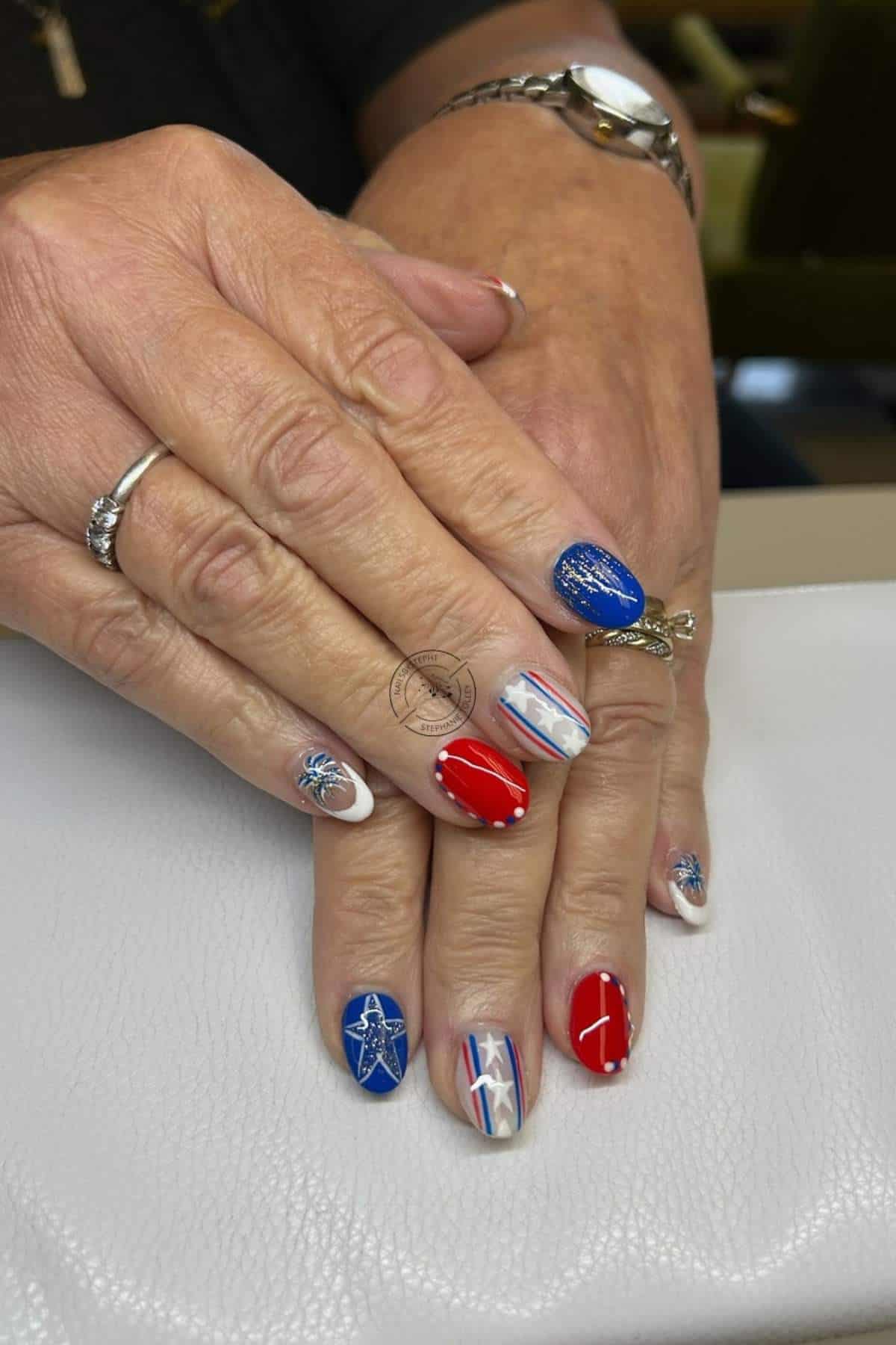 Hands with manicured nails featuring red, white, and blue patriotic designs, including stars, stripes, and glitter, resting on a white surface.