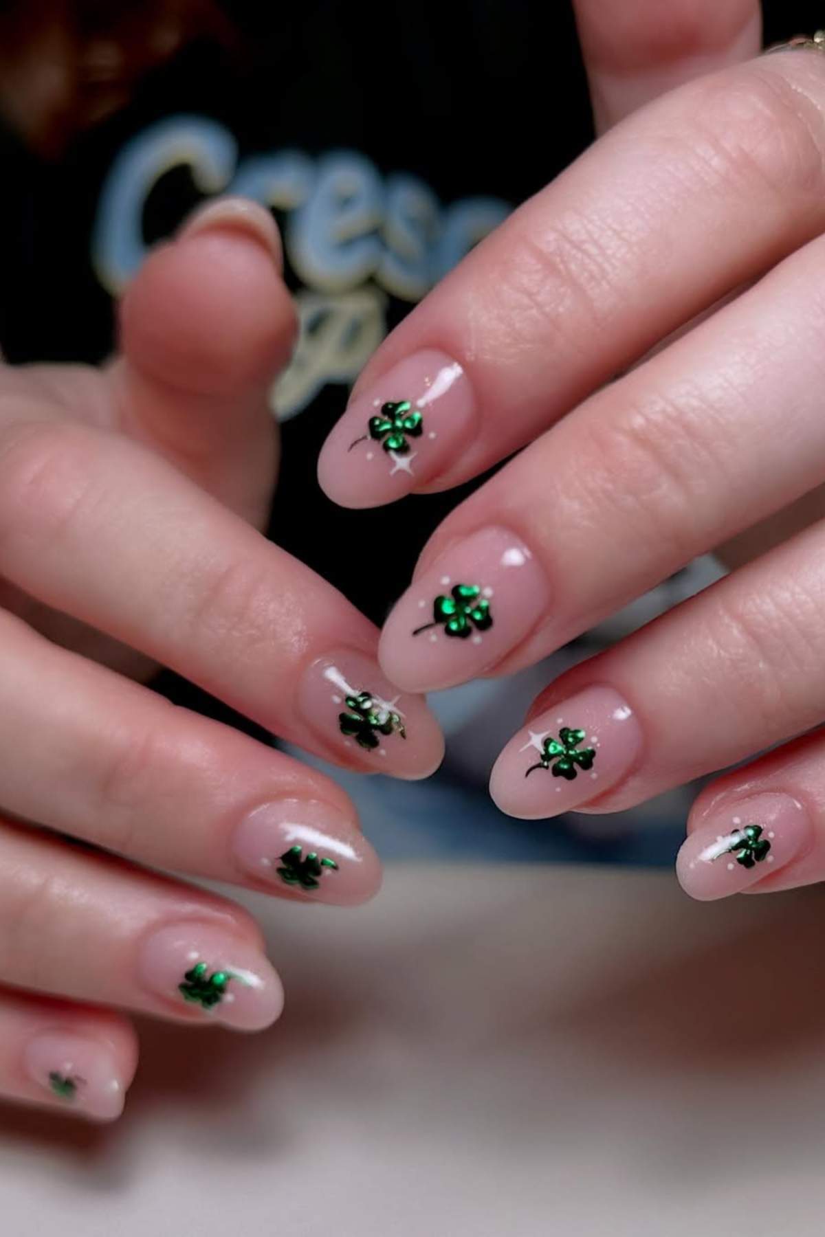 A close-up of hands with nude manicured nails featuring small green four-leaf clover designs on each nail.