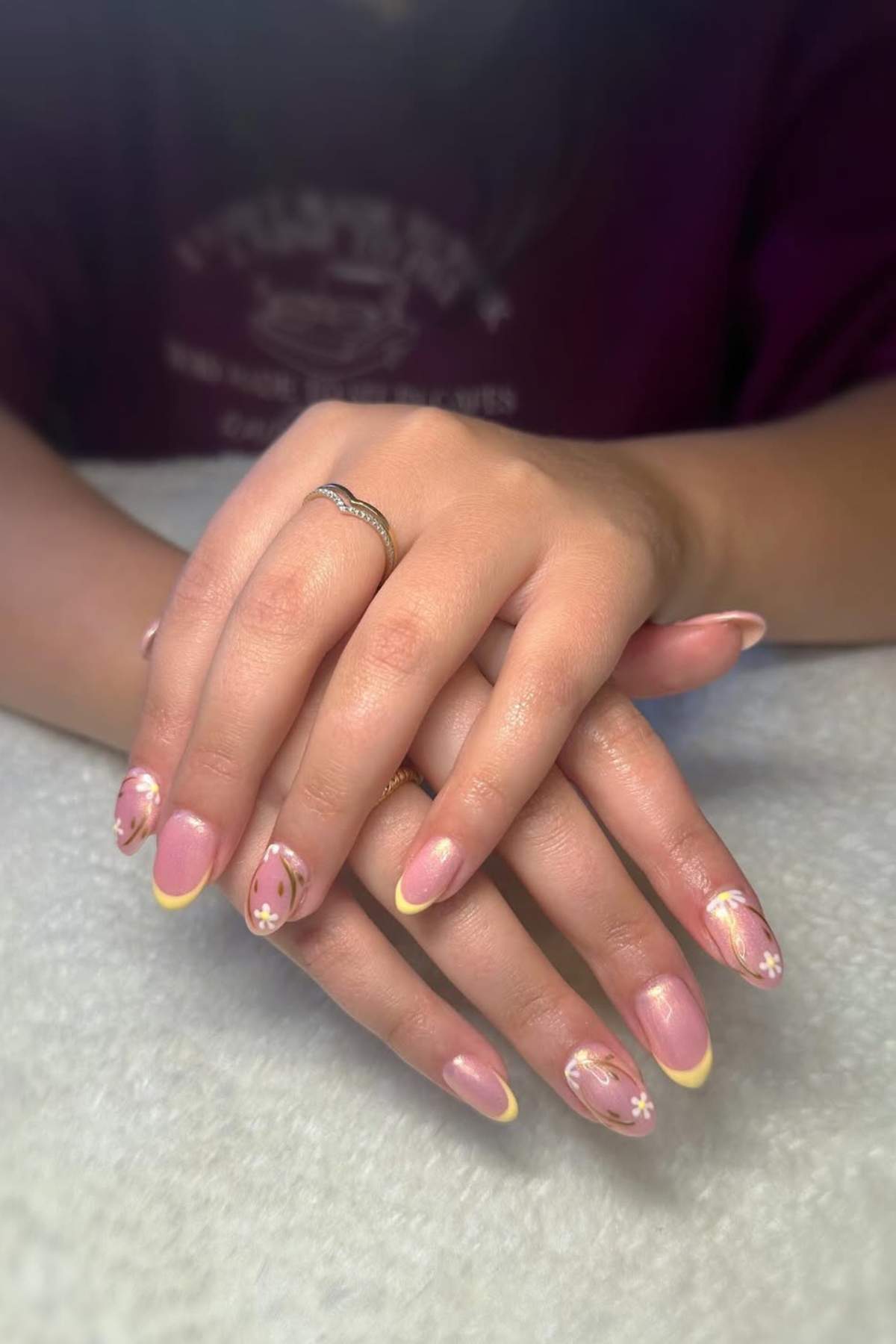 Hands with a silver ring display manicured nails featuring yellow tips and white daisy flower designs on a light pink base, resting on a white surface.