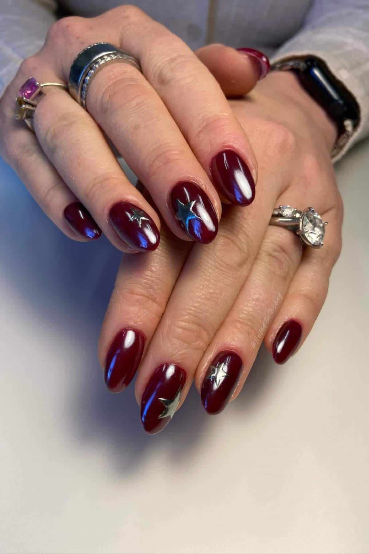 Hands with dark red, almond-shaped nails featuring silver star designs. The person is wearing several rings and a smartwatch on their wrist.