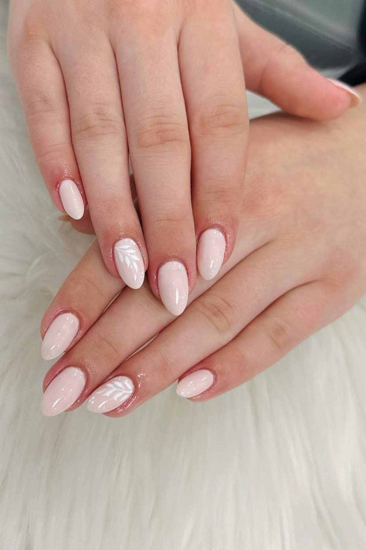 Hands with almond-shaped nails painted light pink; one nail features a white leaf design. The hands are resting on a white, textured surface.