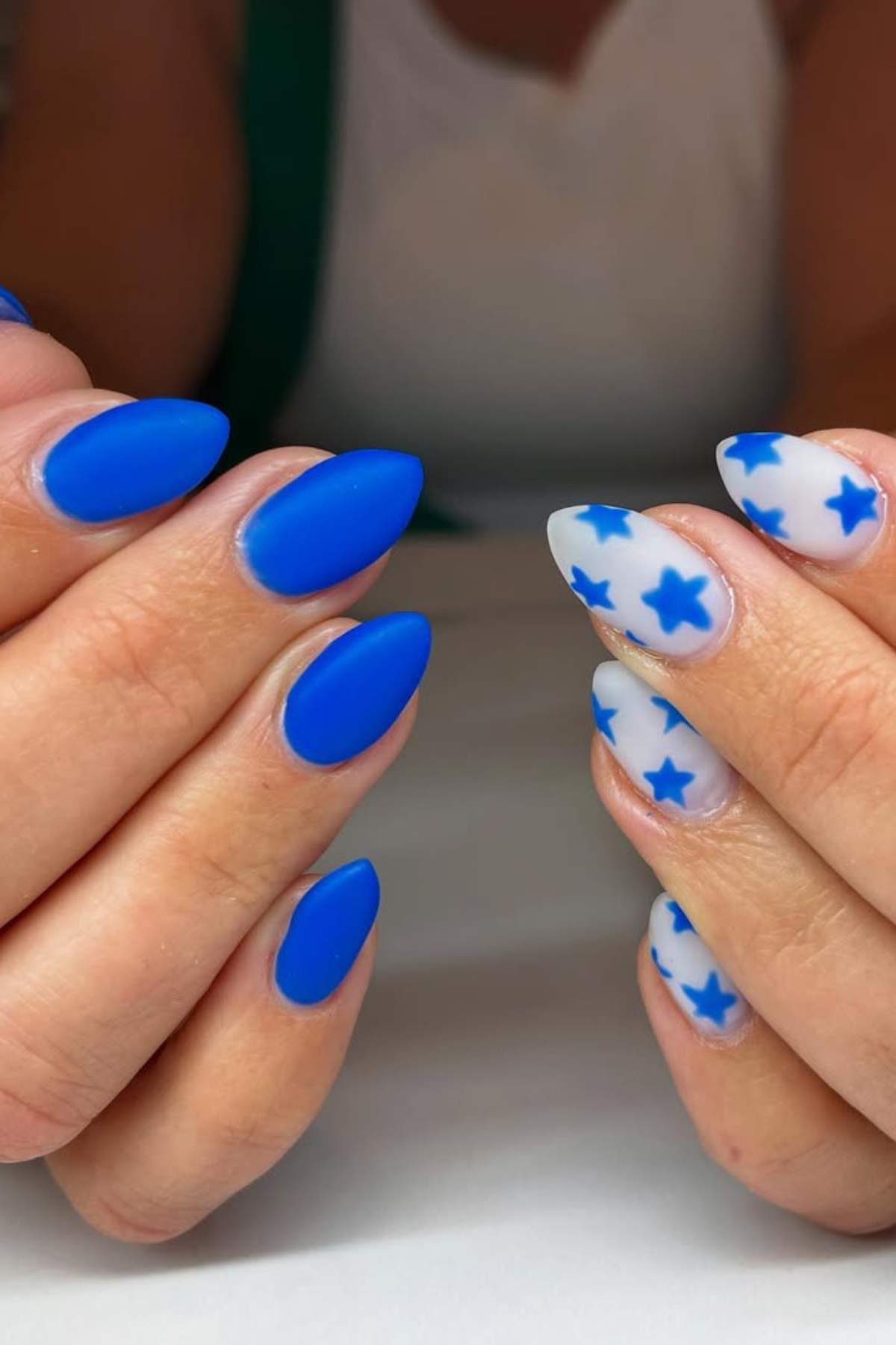 Close-up of hands with almond-shaped nails; one hand has solid matte blue nails, the other has white nails with blue star designs.