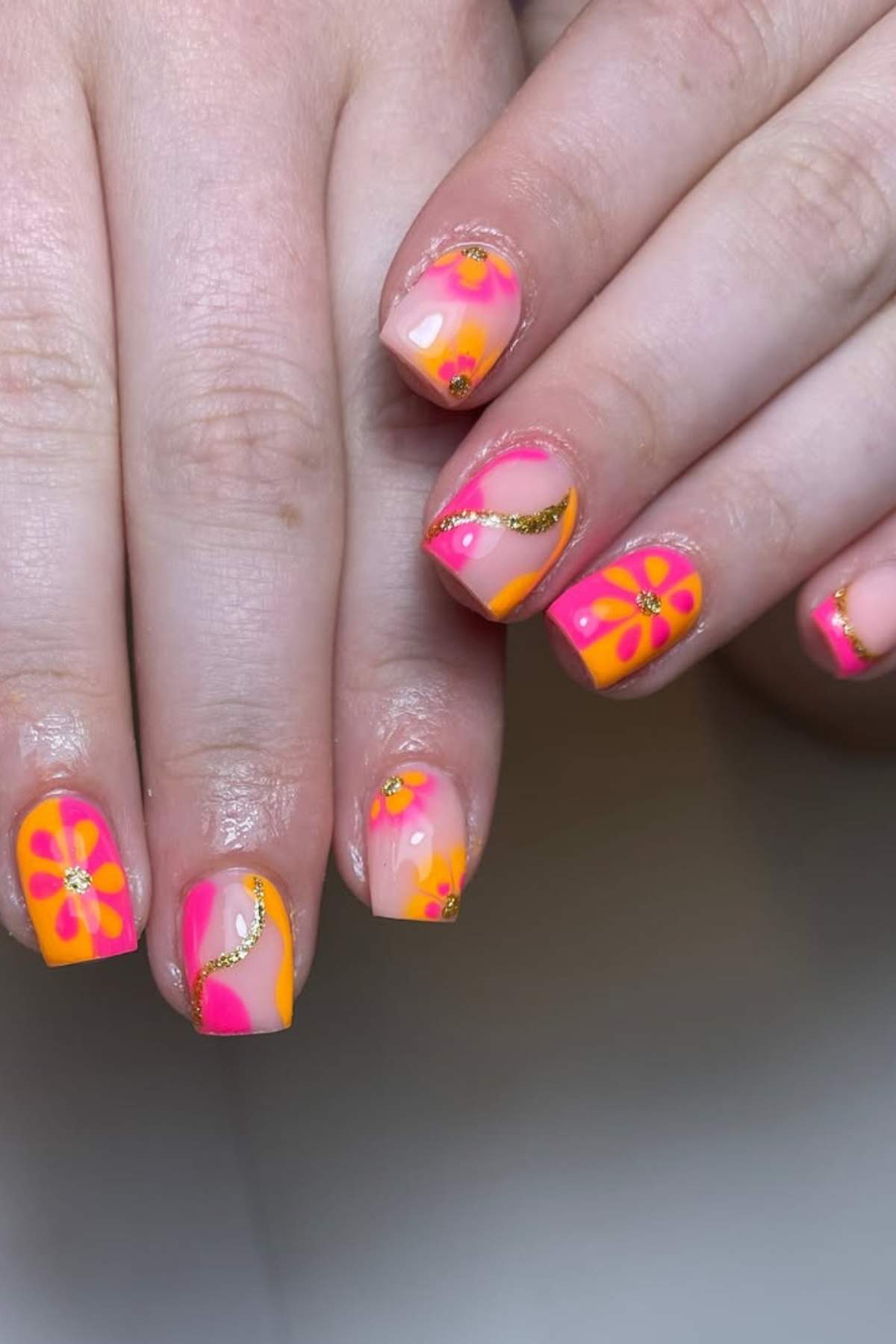 Hands with manicured nails featuring bright pink, orange, and yellow floral designs, gold glitter accents, and a glossy finish.