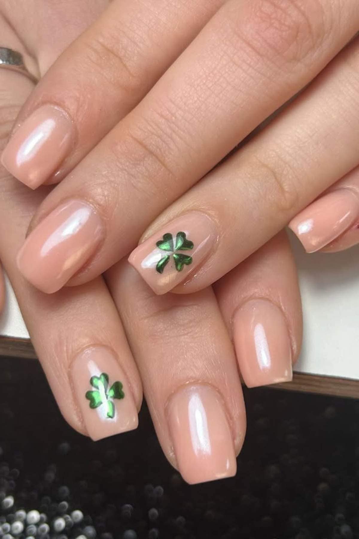Hands with short, square-shaped nude nails; two nails feature small green shamrock designs as accents.