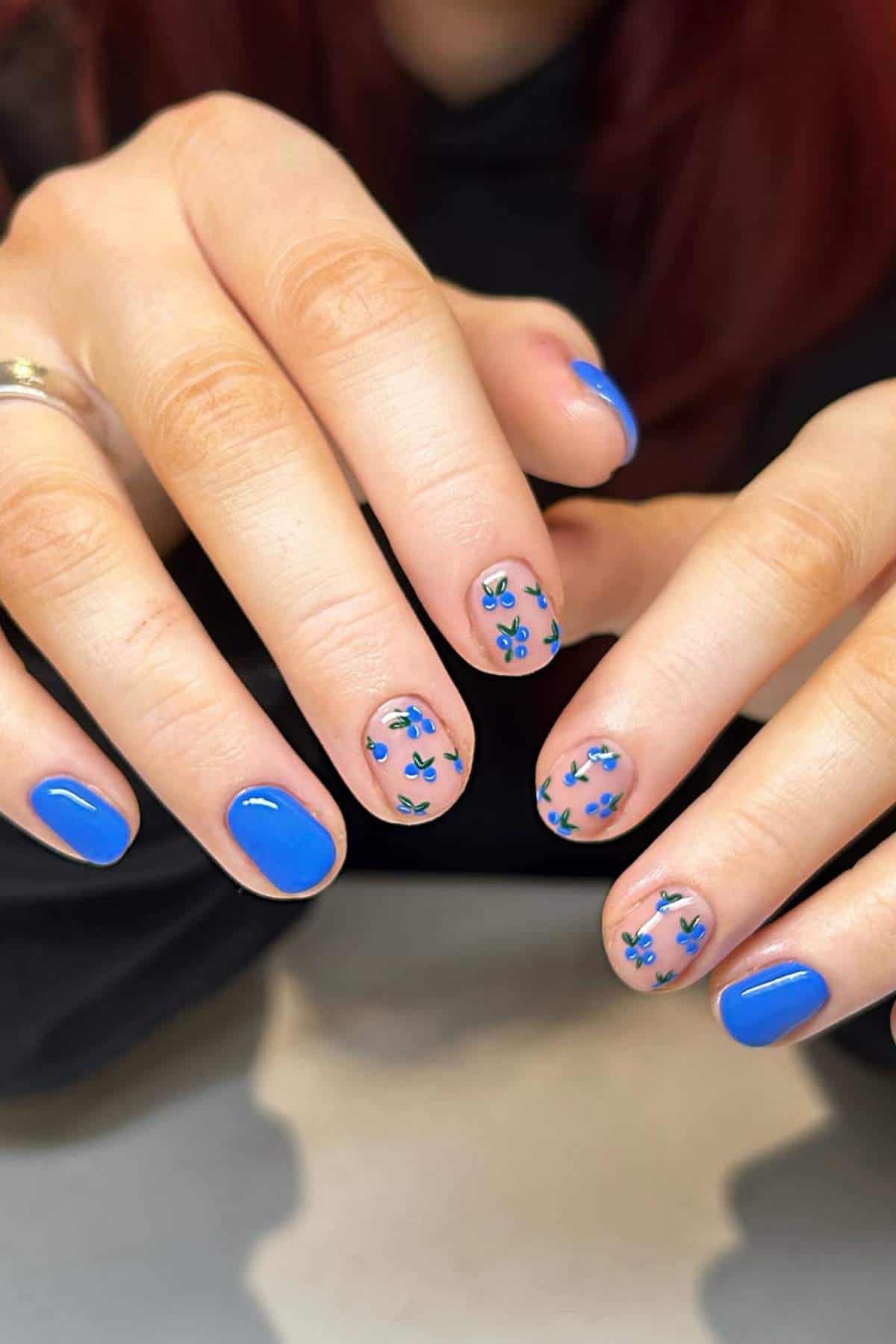 Close-up of hands with short, round nails painted blue; some nails feature small blue flower designs on a clear base.