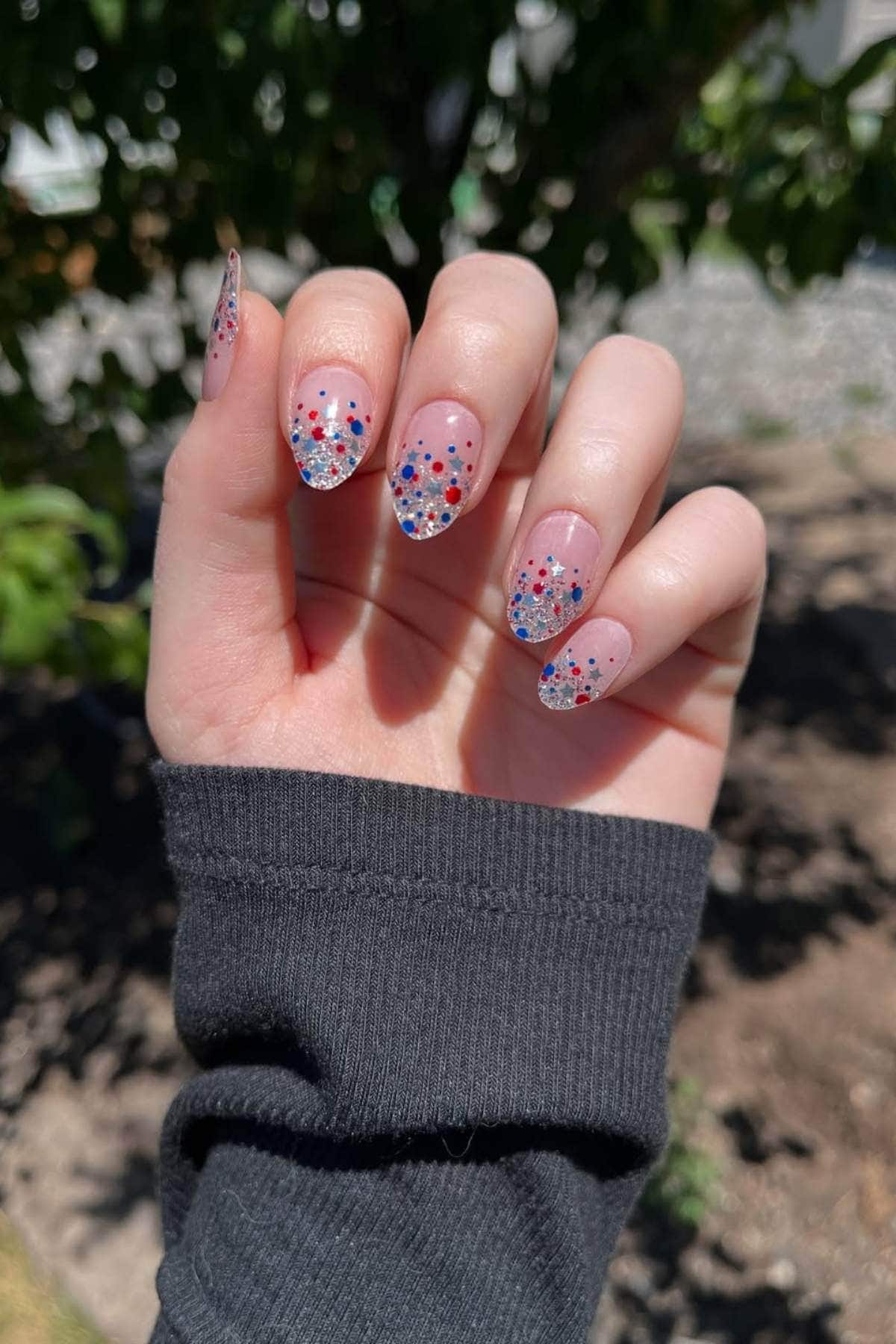 A hand with almond-shaped nails featuring clear tips decorated with red, blue, and silver glitter dots, held against an outdoor background with greenery.