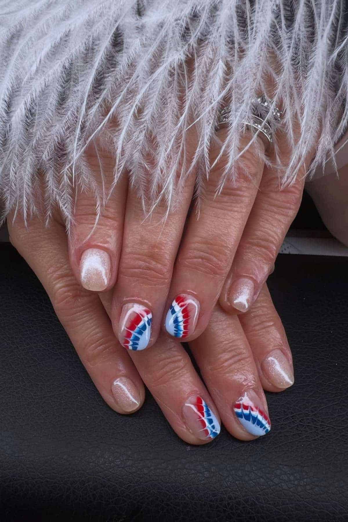 A pair of hands with manicured nails; some nails are painted white, while others have a red, white, and blue zigzag pattern. A white feathery fabric rests over the hands.