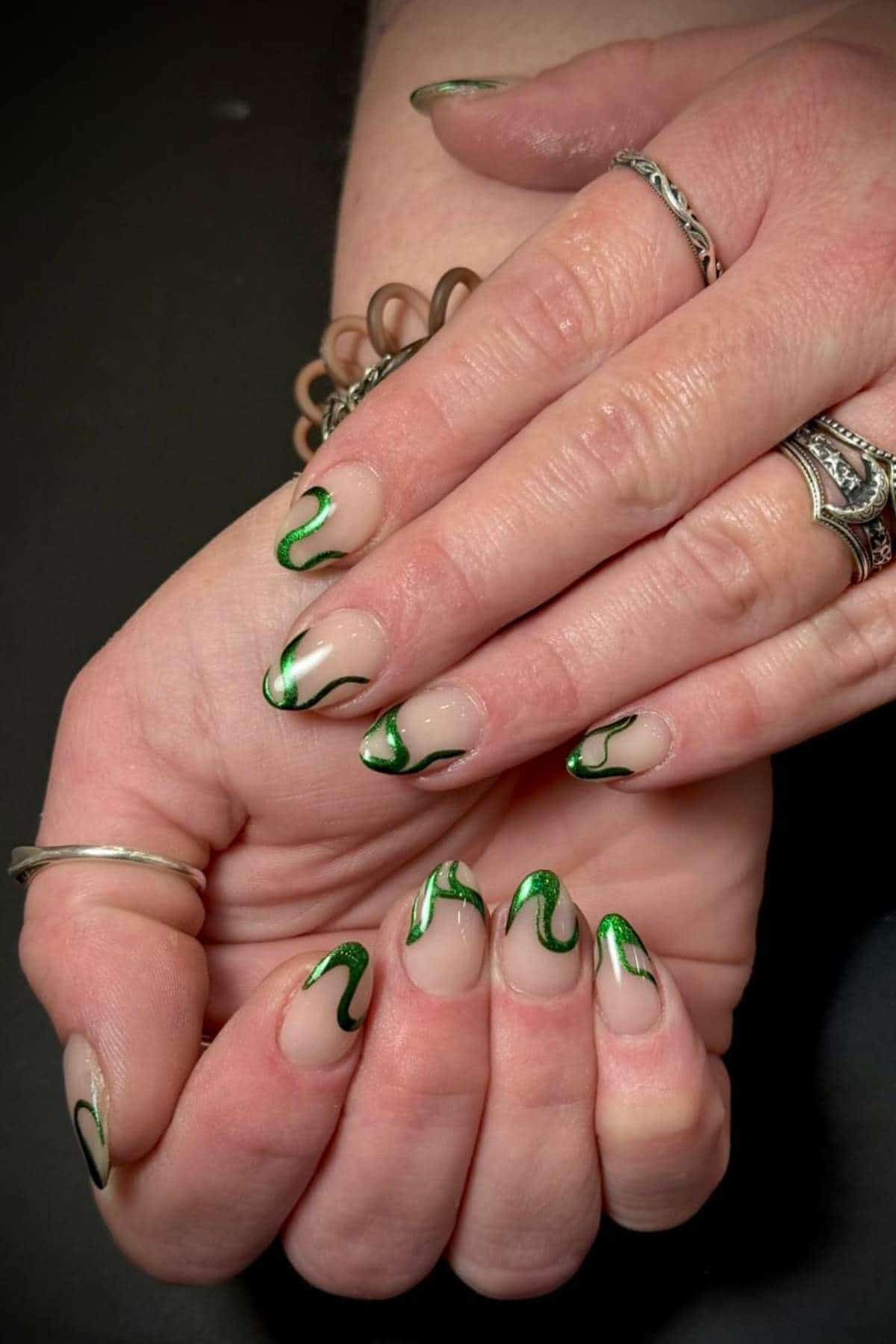 Hands with almond-shaped nails featuring a clear base and green abstract swirl designs. The person is wearing silver rings and a brown spiral hair tie.