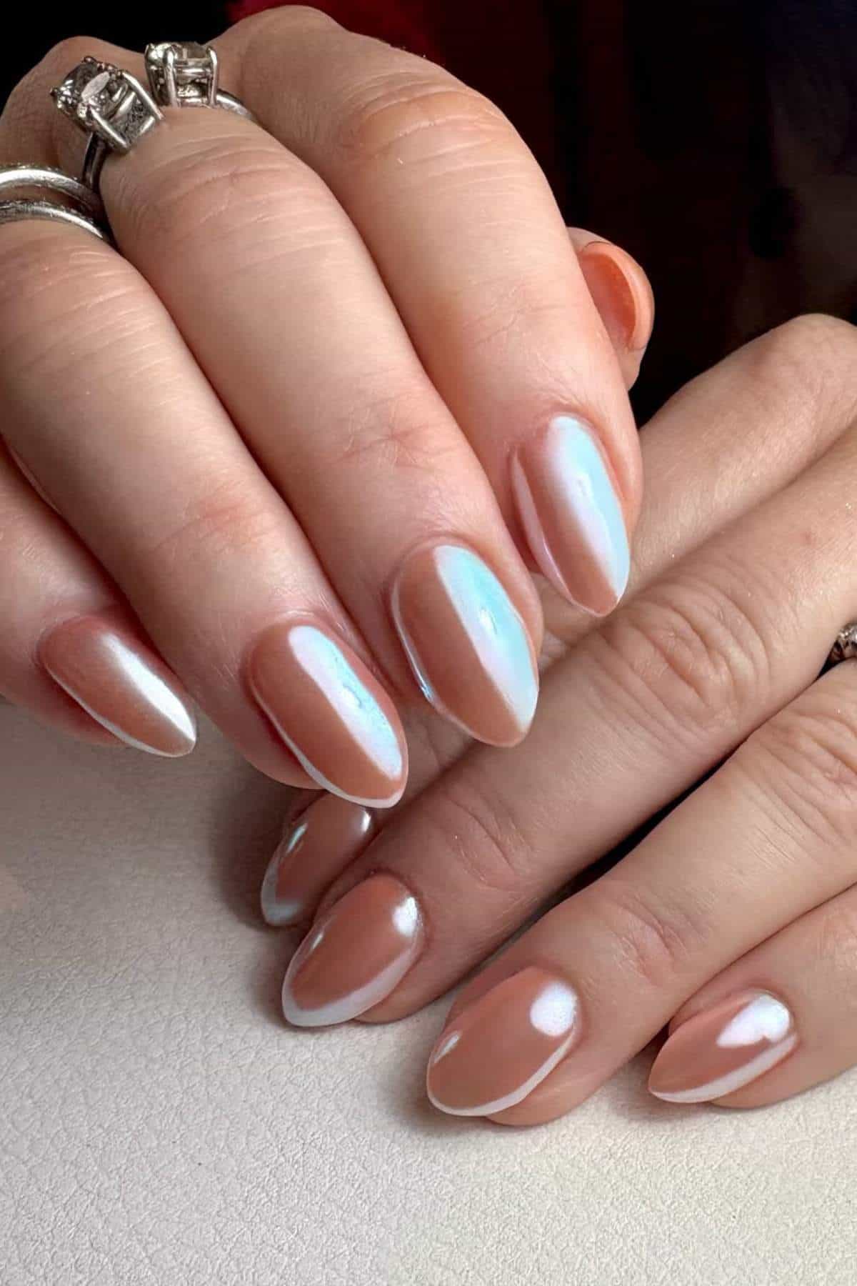 Close-up of hands with almond-shaped nails featuring a pearly, iridescent white manicure. The person is wearing silver rings.
