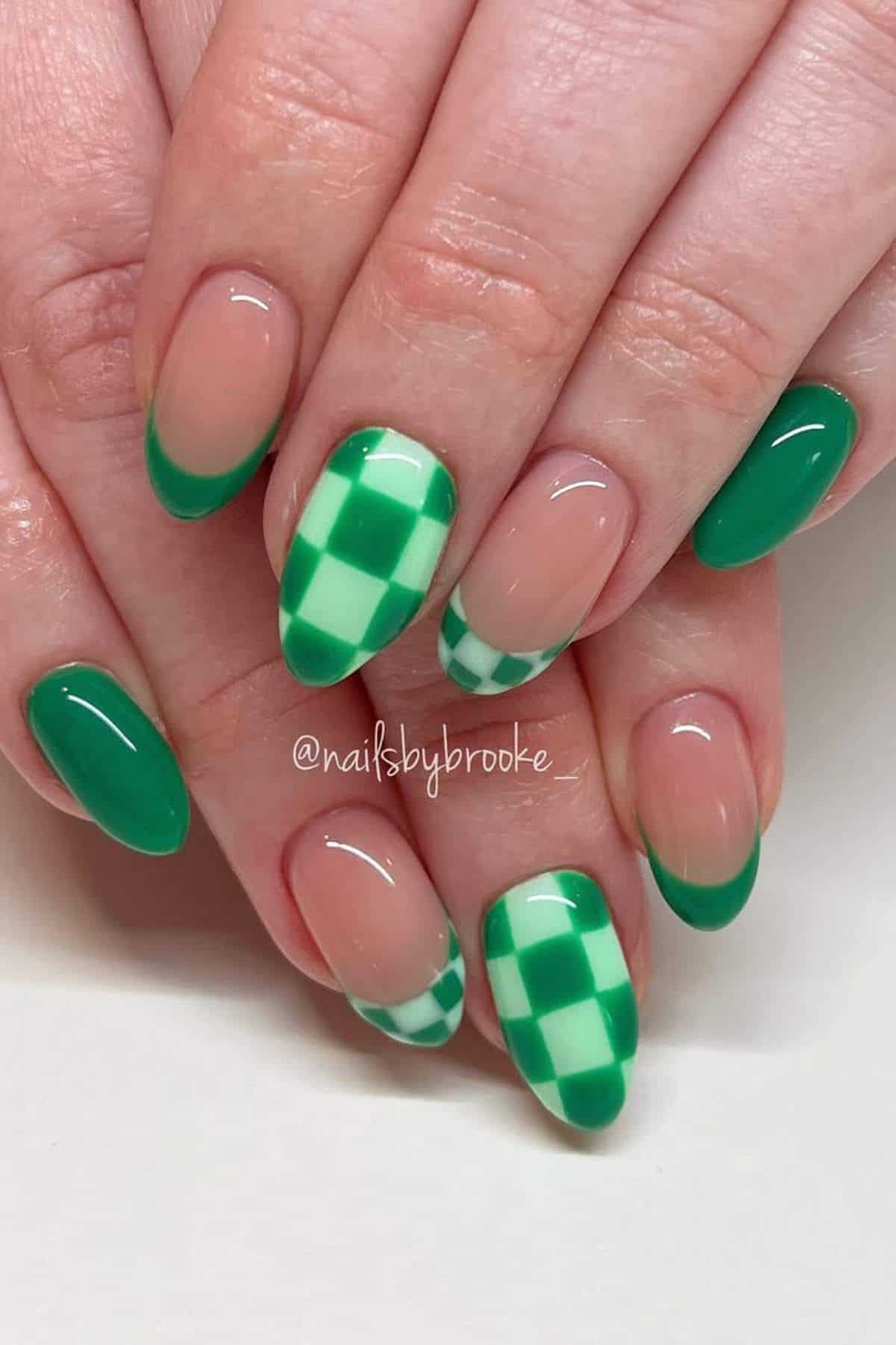 Hands with almond-shaped nails featuring a green French tip design and green and white checkered pattern on two accent nails.