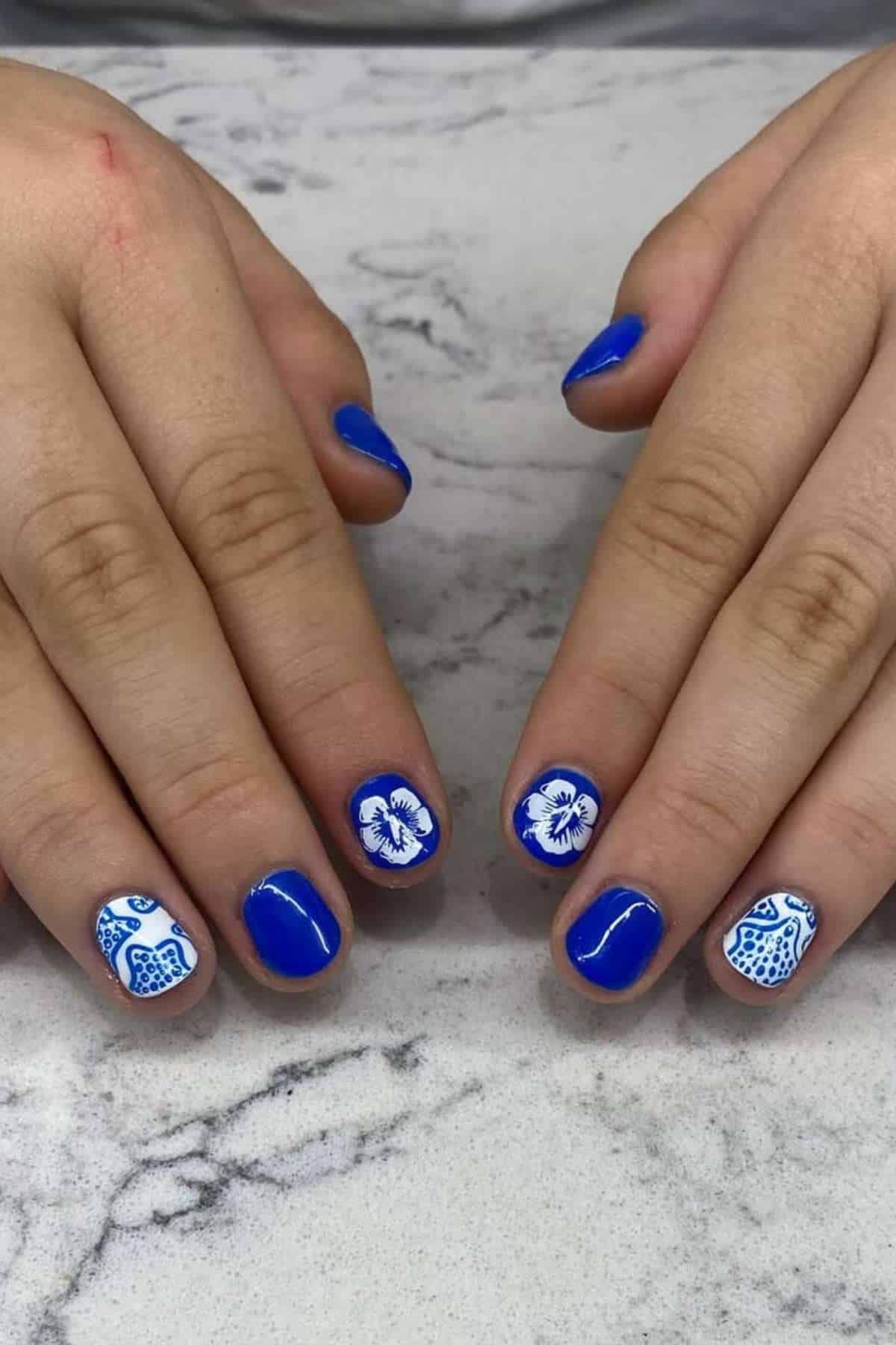 Two hands with short nails painted blue, white accent nails featuring blue floral and abstract designs, resting on a light marbled surface.