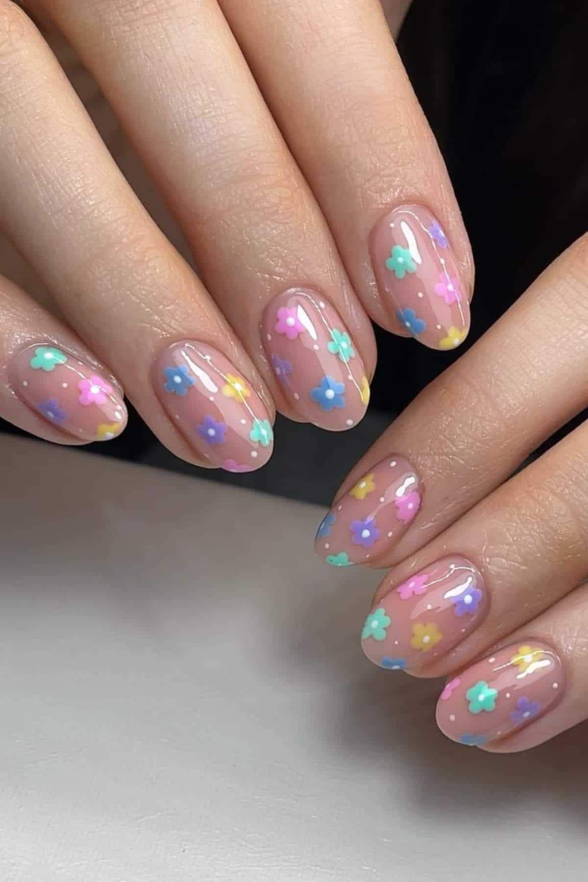 Hands with oval-shaped nails featuring a clear base and small, colorful flower designs in pastel shades on each nail.