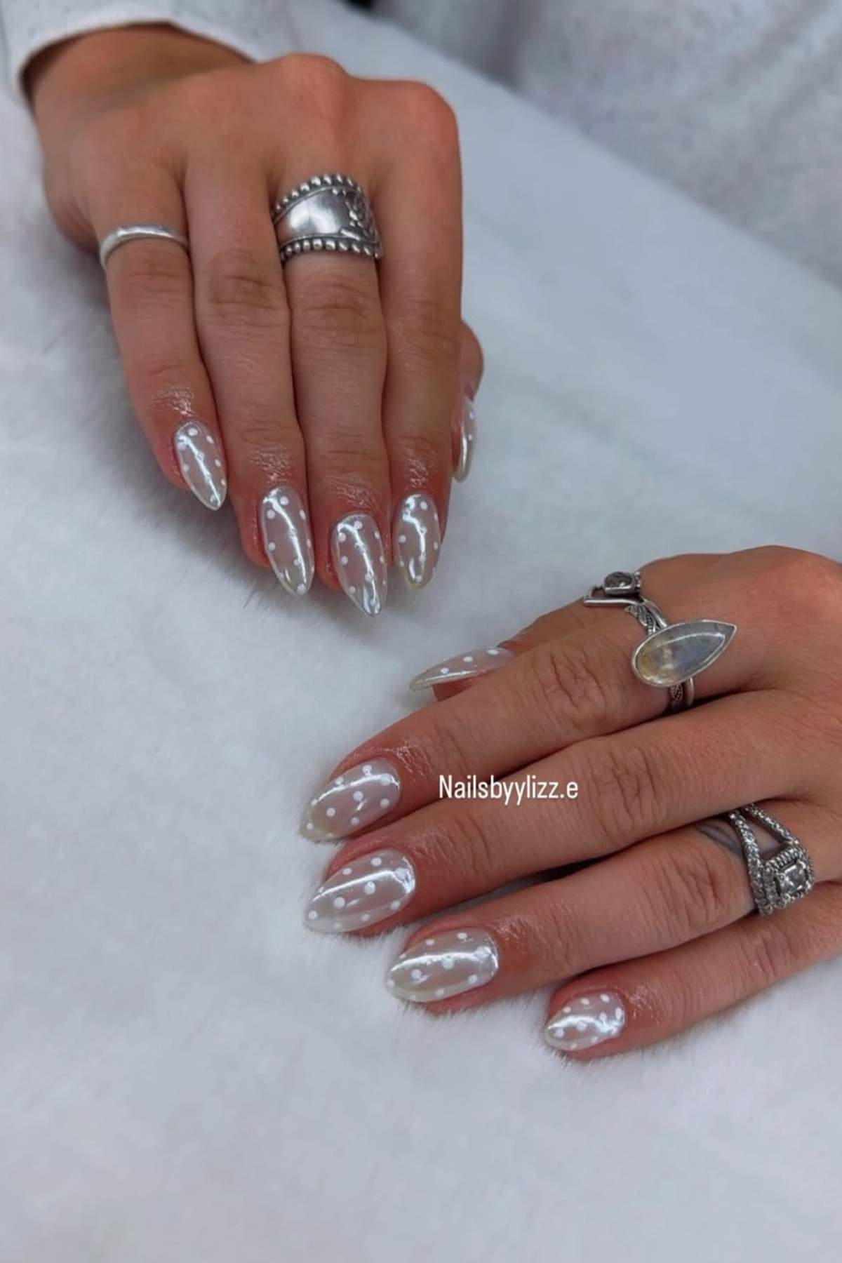 Two hands with almond-shaped nails featuring a milky white base and glossy, cloud-like designs, adorned with multiple silver rings on each hand.