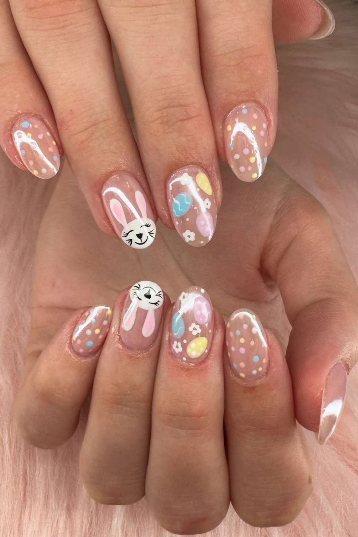 Hands with pastel Easter-themed nail art, featuring bunny faces, eggs, hearts, dots, and florals on a clear base.
