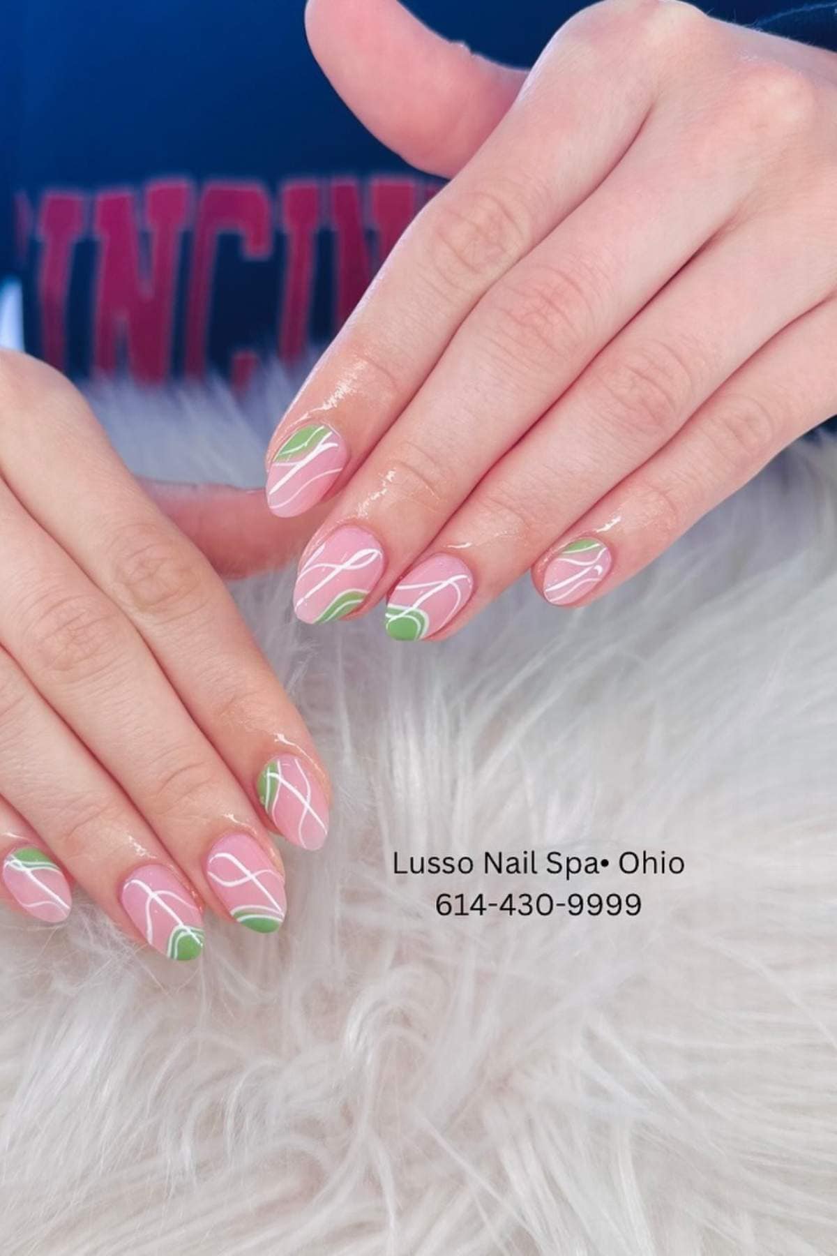Hands with almond-shaped nails featuring pink polish, white and green abstract line designs. "Lusso Nail Spa • Ohio 614-430-9999" text appears on a furry white surface.