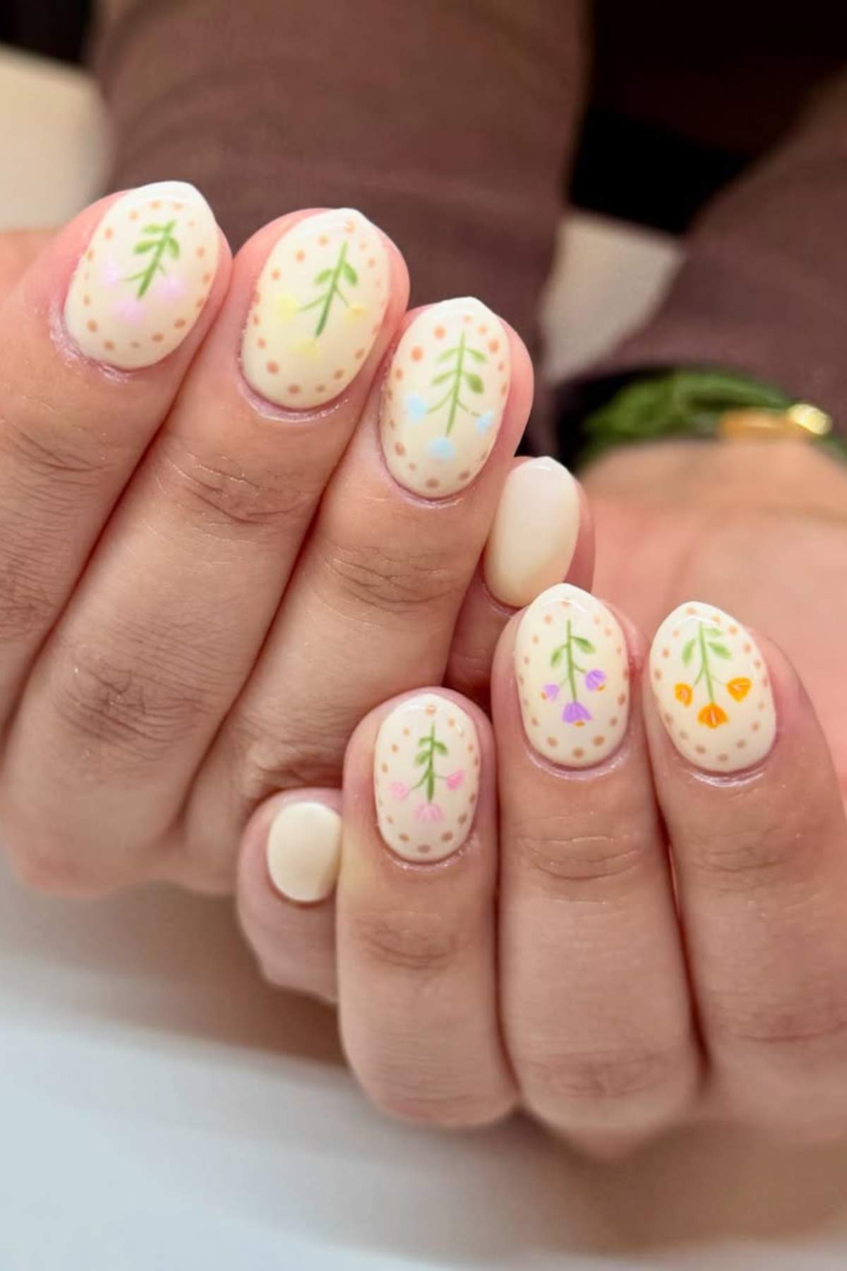 Close-up of hands with short, oval-shaped nails painted off-white and decorated with simple, colorful leaf and dot patterns.