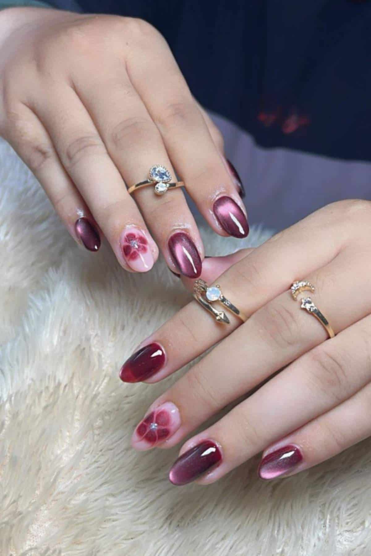 Two hands with almond-shaped nails painted in burgundy with a glossy finish; two accent nails feature pink backgrounds with hand-painted floral designs. Multiple gold rings are worn on the fingers.