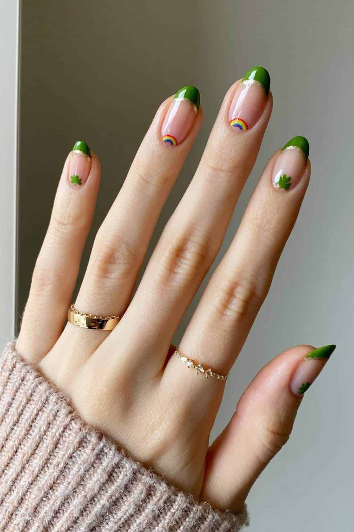 A hand with green French tip manicure, small shamrock and rainbow designs on the nails, wearing two rings and a beige knit sweater.