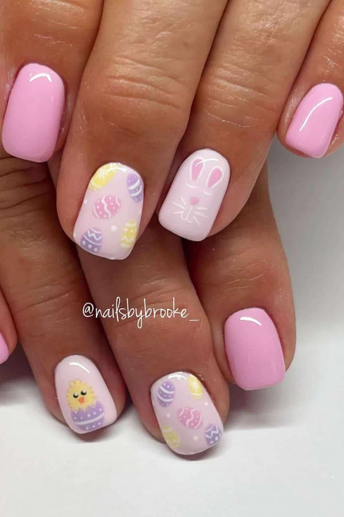 Hands with short, rounded pink nails. Two accent nails feature Easter-themed designs, including decorated eggs, a bunny face, and a chick in a shell.