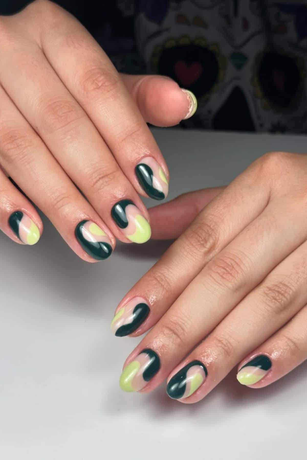 Hands with oval-shaped nails featuring abstract green and yellow nail art, displayed against a white surface.