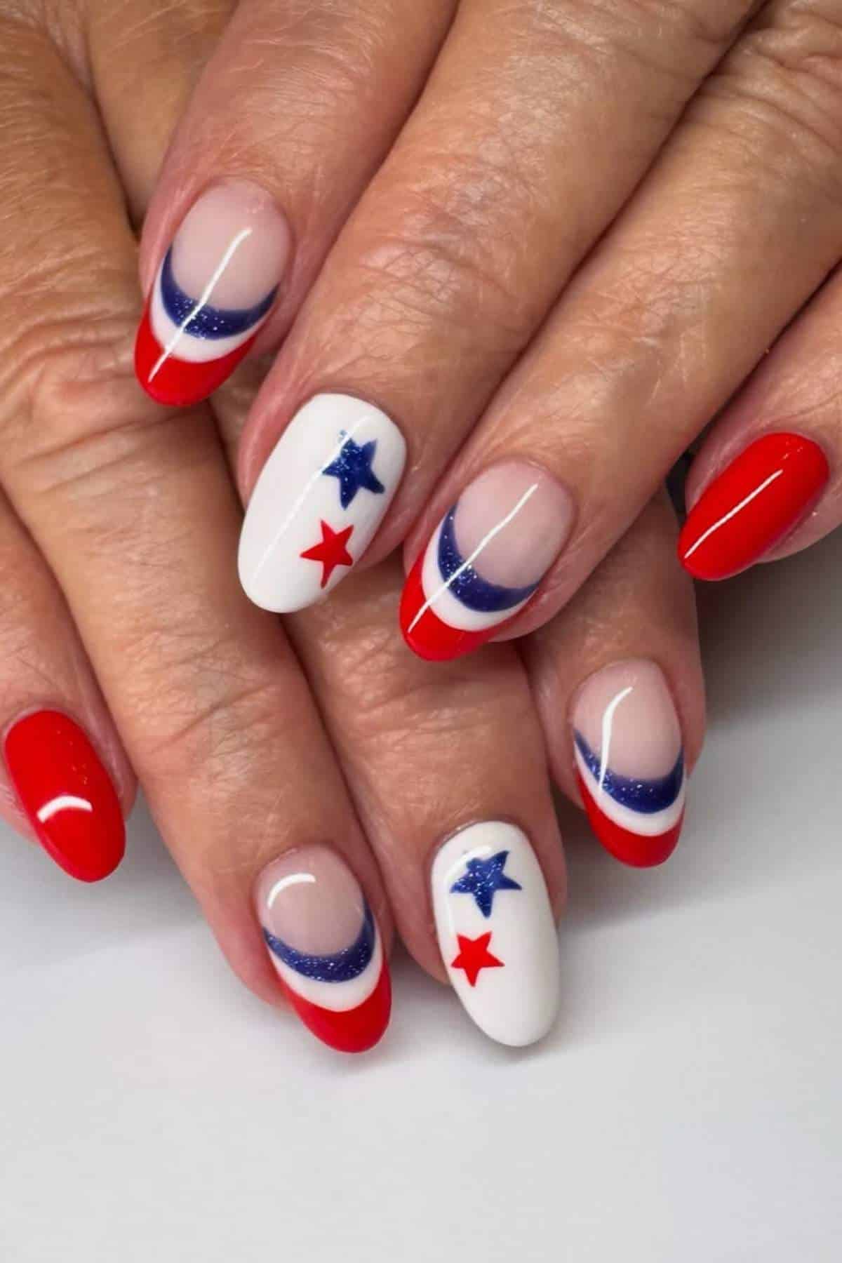 Hands with manicured nails featuring red tips, blue glitter accents, and white nails decorated with red and blue star designs.