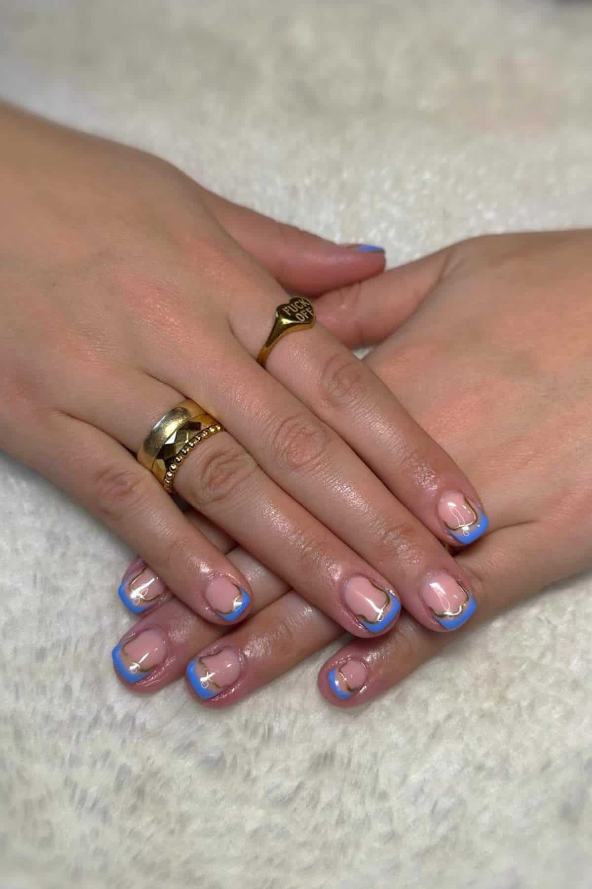 Two hands with gold rings rest on a white surface, showing a manicure with blue and gold French tips on natural, rounded nails.