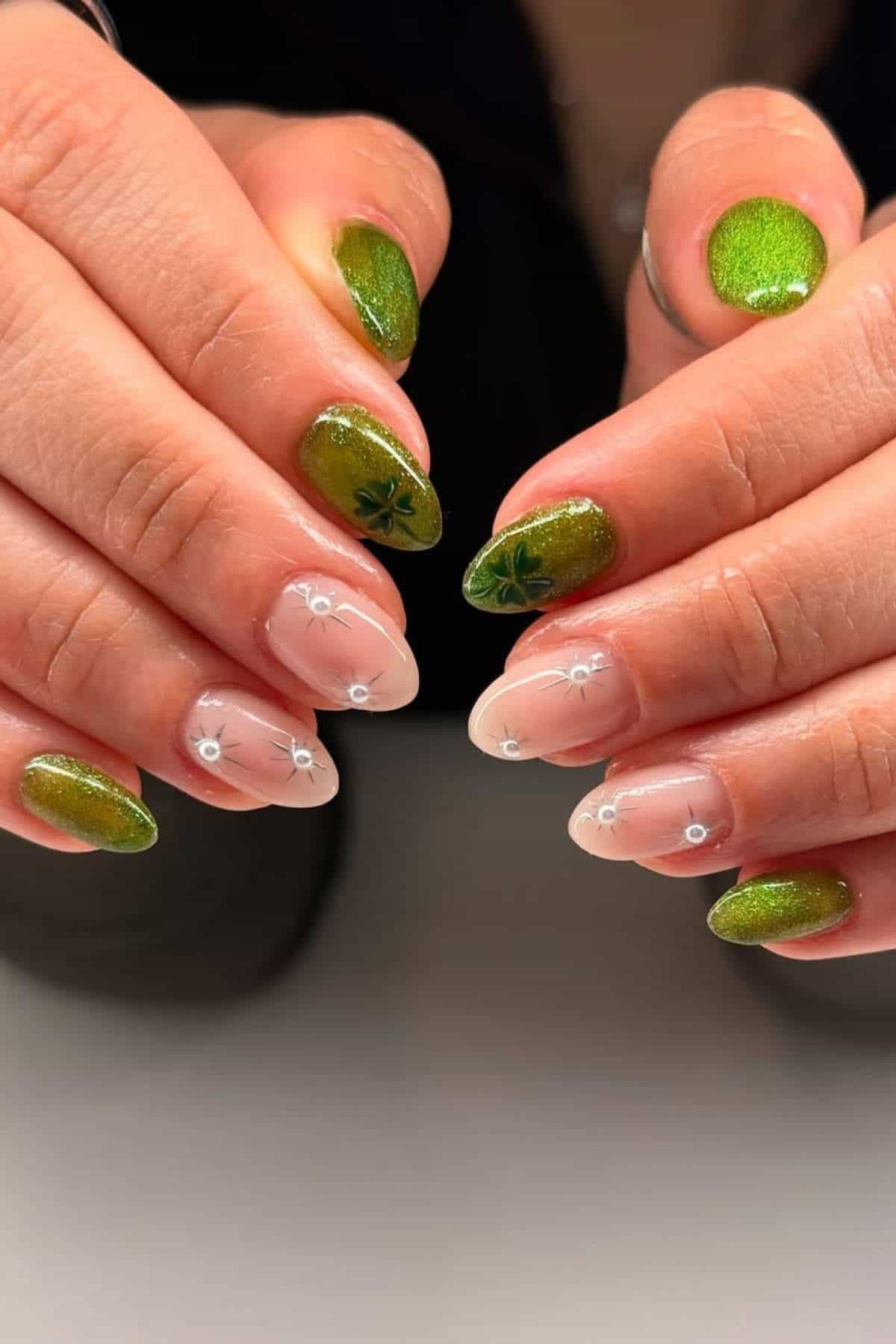Hands with almond-shaped nails featuring a green shimmery polish, some with small leaf designs, and others with a nude base and simple white flower accents.