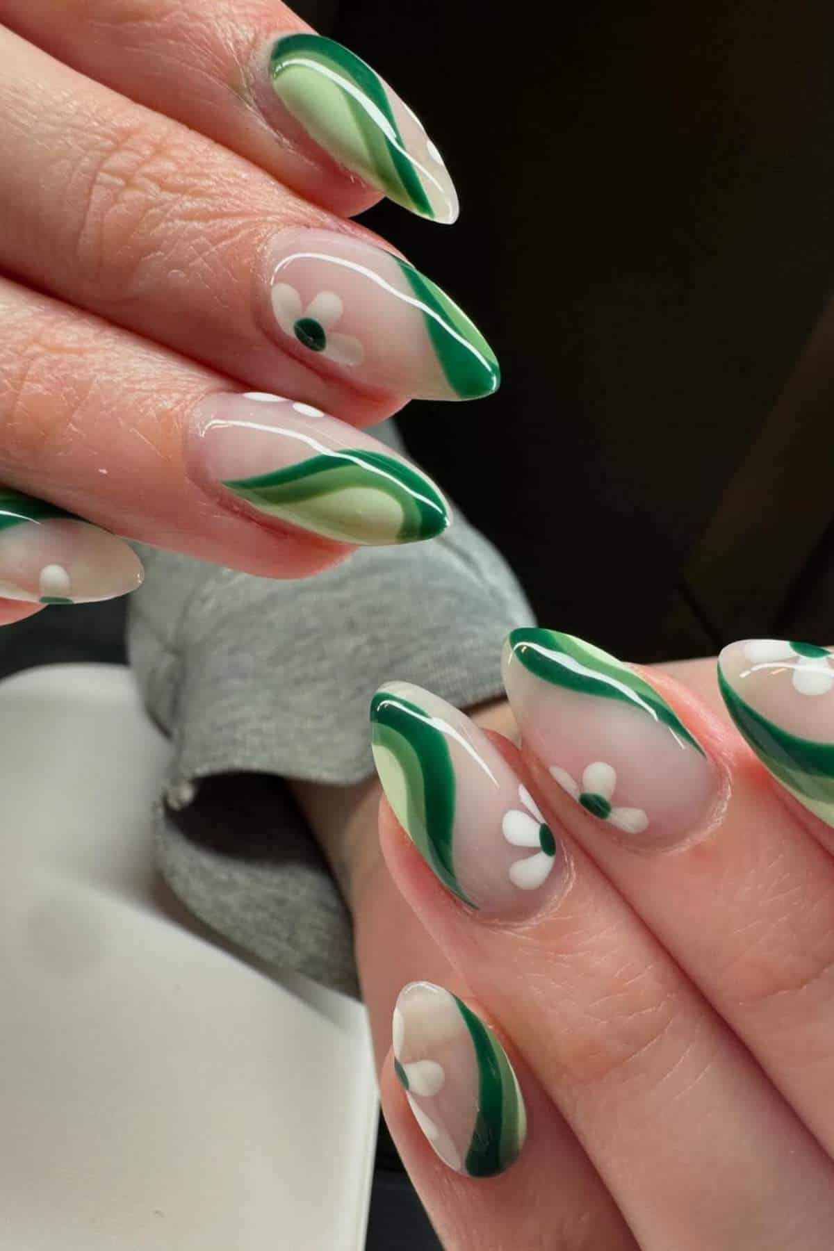 Close-up of hands with almond-shaped nails featuring green abstract lines and white daisy flower designs on a clear base.