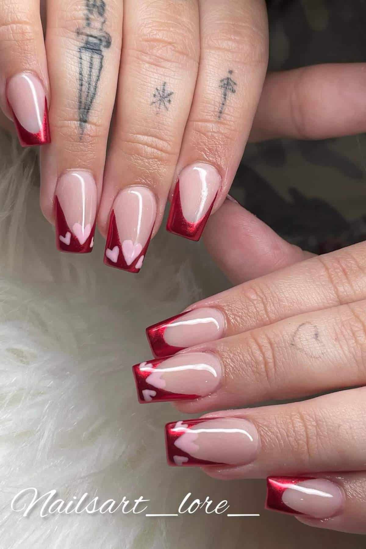 Hands with square-shaped nails featuring a red French tip design and small pink heart accents, shown against a white background. Some fingers have visible tattoos.