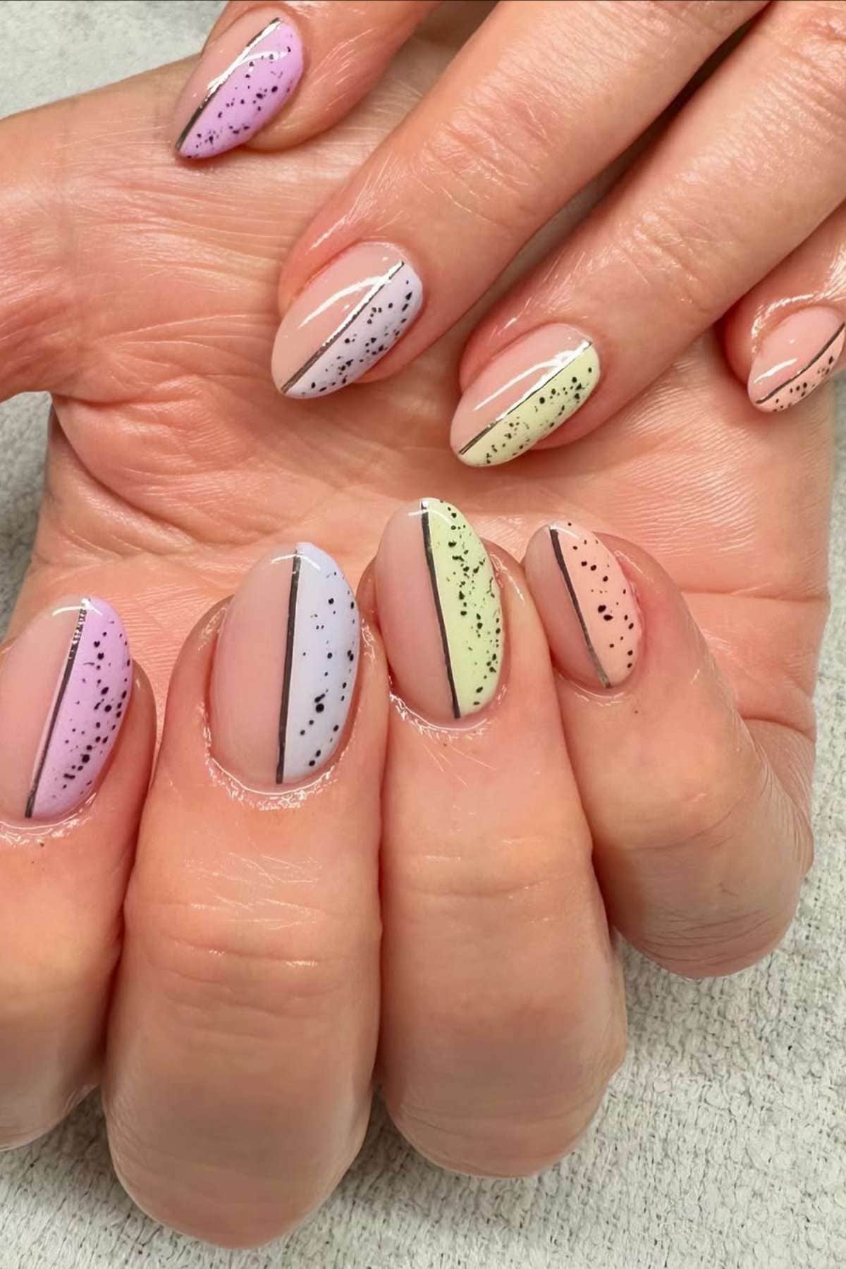 Hands with manicured nails, each nail featuring a diagonal split design in pastel colors with black speckles and a thin black line dividing the sections.