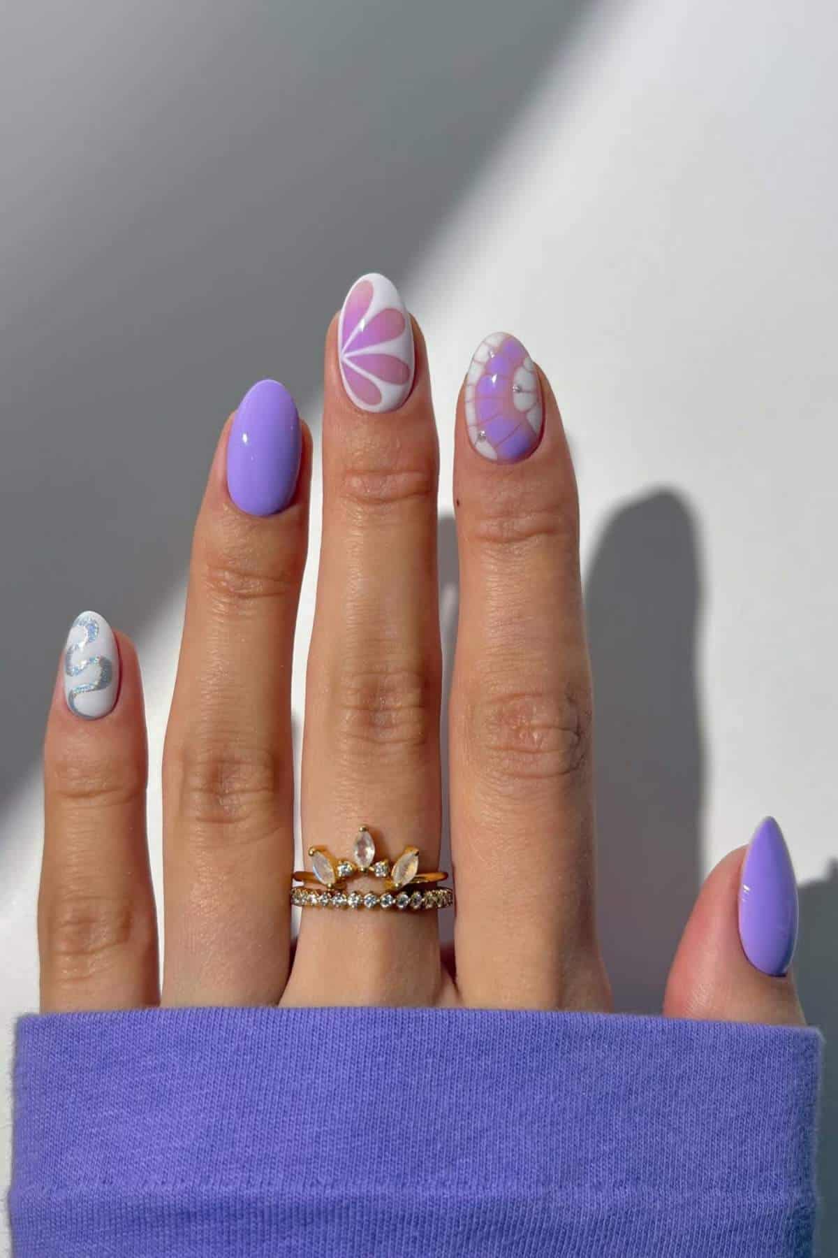 A hand with lilac and white flower-themed nail art, wearing gold rings, rests against a light background.