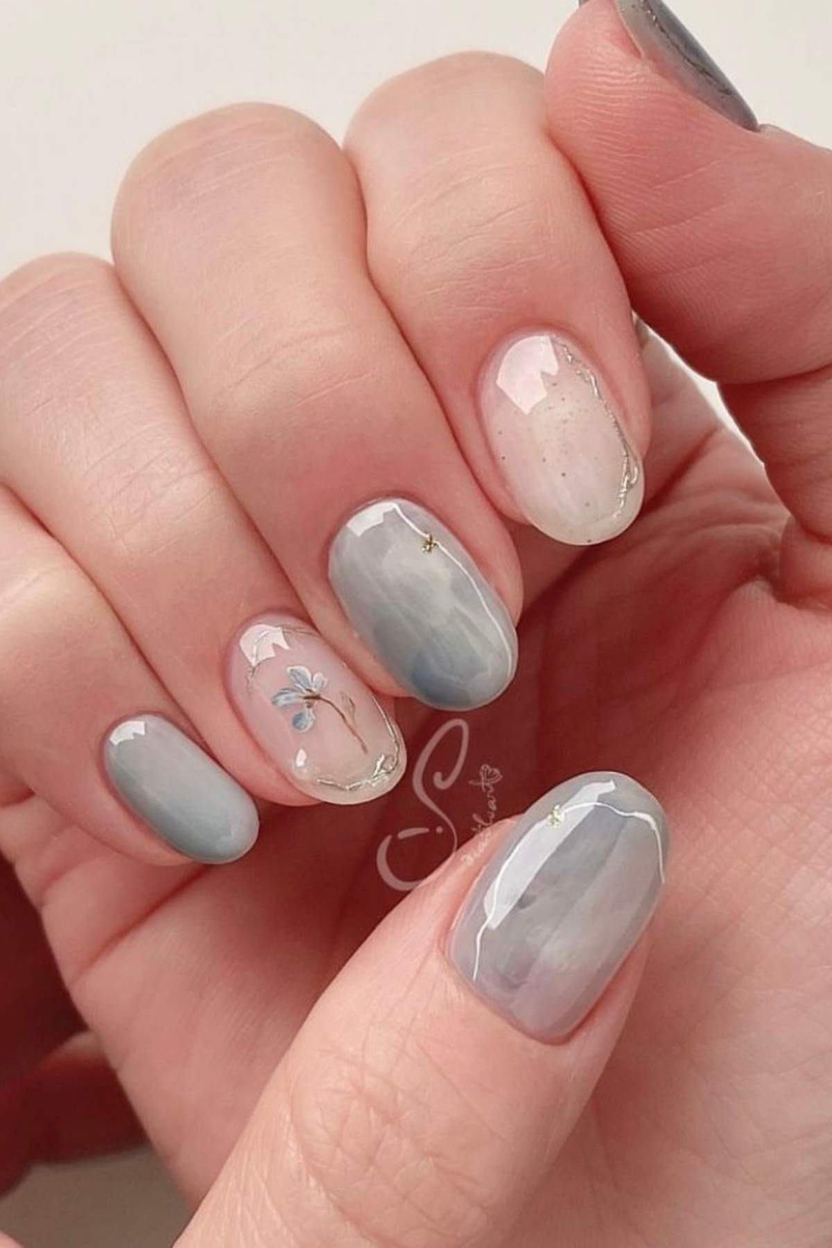 A hand with medium-length, oval-shaped nails painted in soft, cloudy gray and white tones, some featuring delicate floral and gold accent details.
