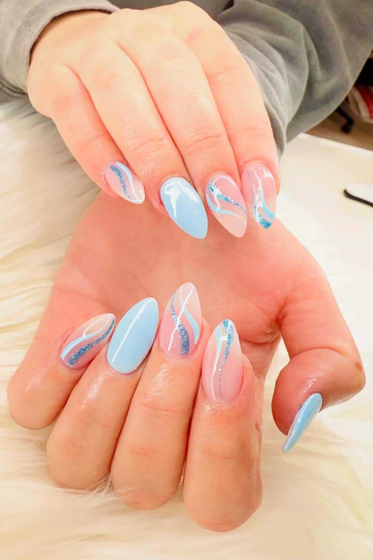 Hands with almond-shaped nails painted in light blue and nude tones, decorated with blue and white wavy lines and glitter accents.