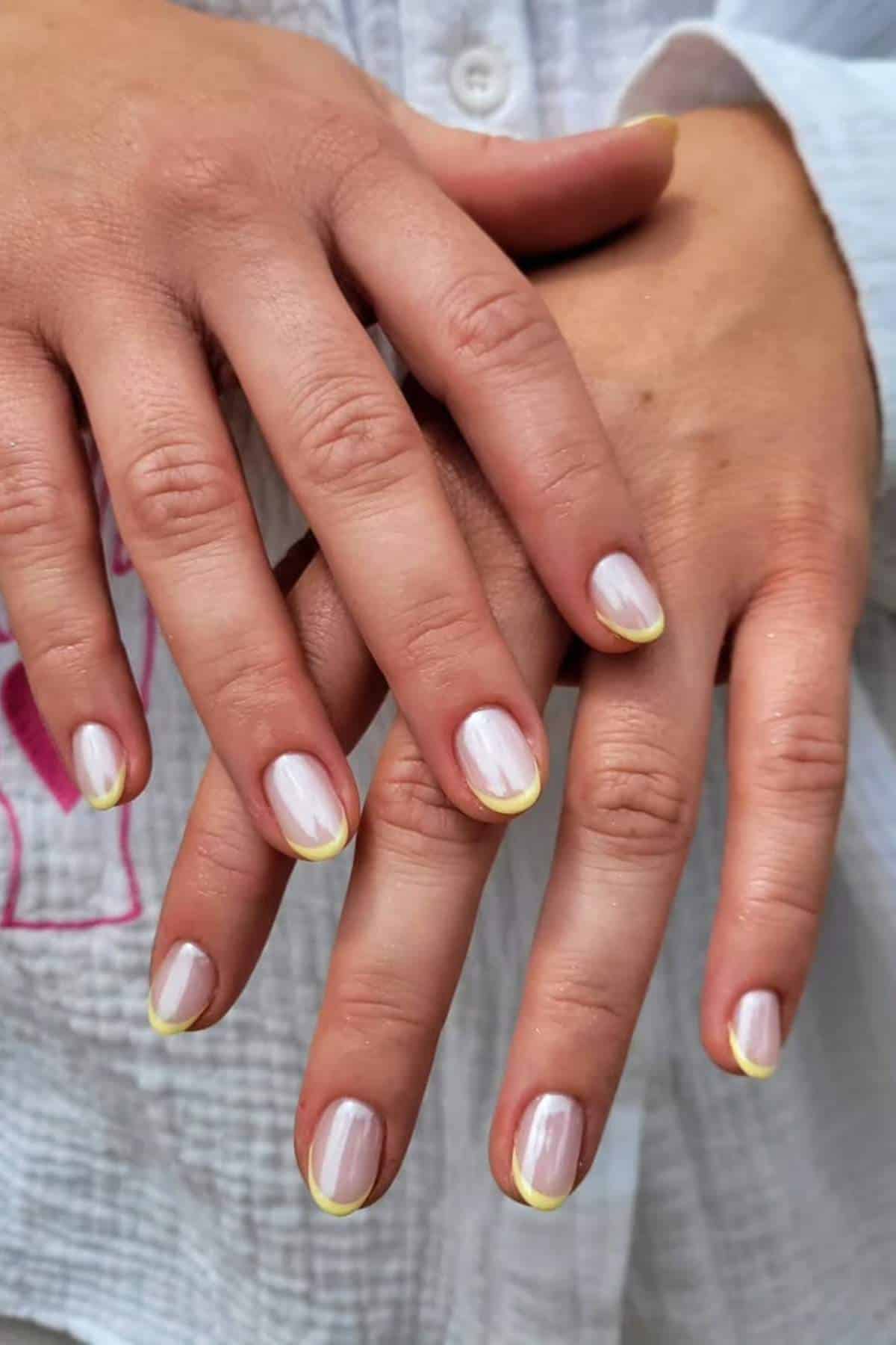 Two hands with short, oval-shaped nails painted in a pale pink shade, each nail featuring a thin, light yellow French tip.