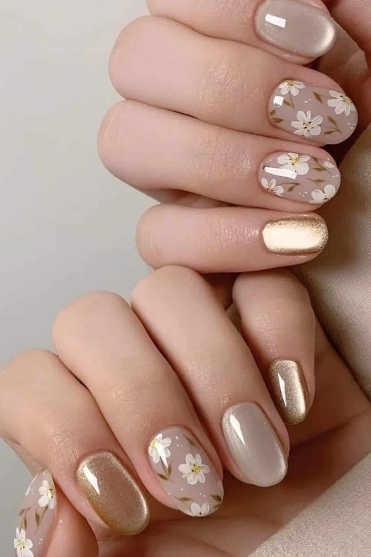 Hands with manicured nails featuring a mix of gold, nude, and floral designs with white flowers and green leaves on a neutral background.