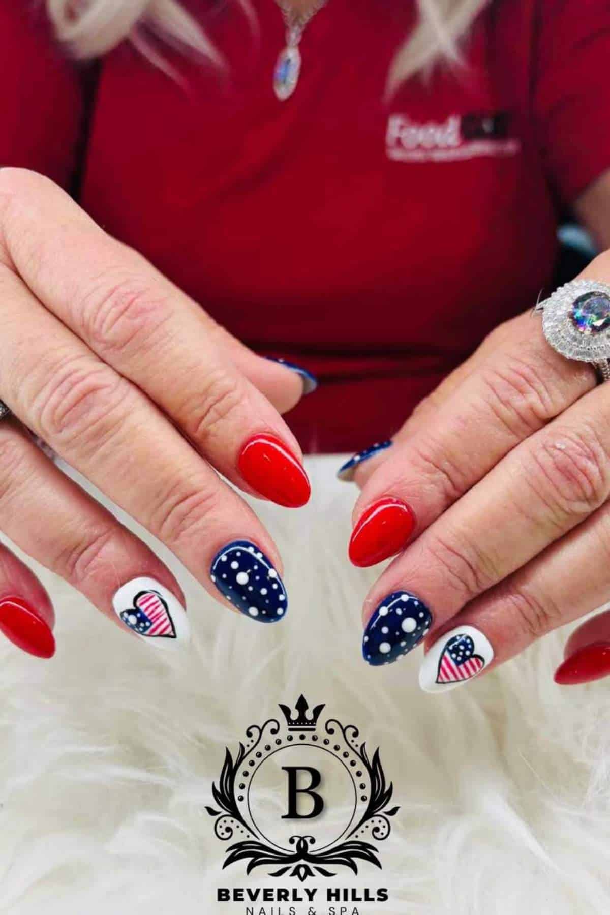 A close-up of hands with manicured nails featuring red, blue with white polka dots, and American flag designs on two accent nails. "Beverly Hills Nails & Spa" logo at the bottom.
