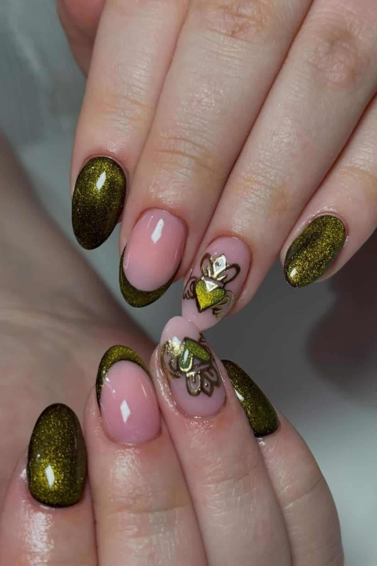 A hand with almond-shaped nails featuring shimmery olive green polish and accent nails with nude pink polish and metallic gold heart and flower designs.