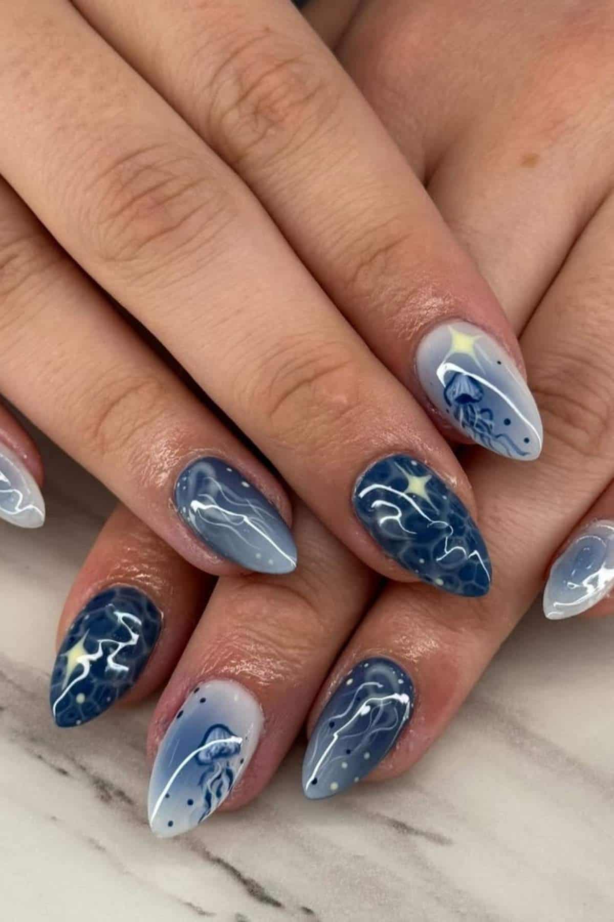 Hands with almond-shaped nails painted in various shades of blue and white, featuring abstract wave and dot designs on a marble surface.