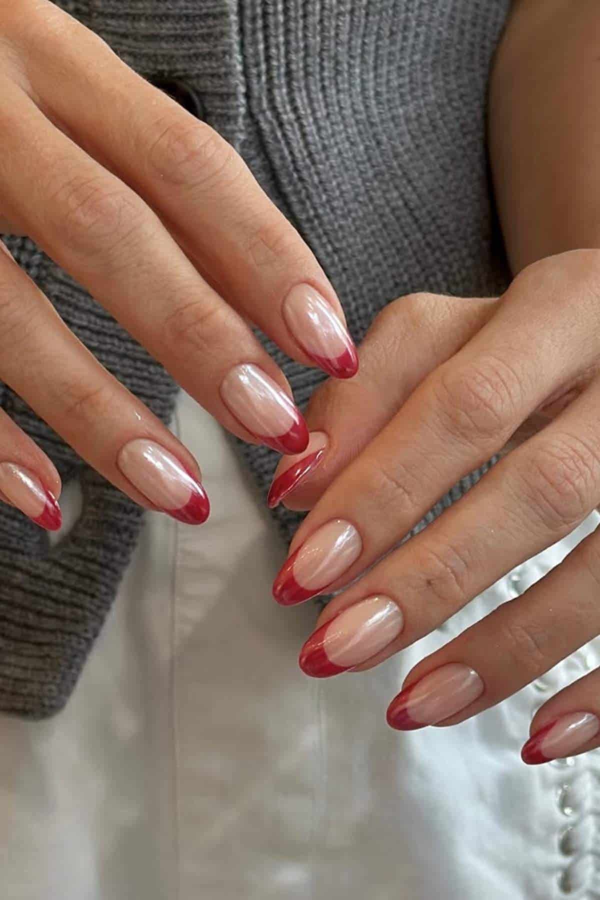 A person’s hands with almond-shaped nails featuring a natural base and glossy red tips, displayed against a gray knit and white fabric background.