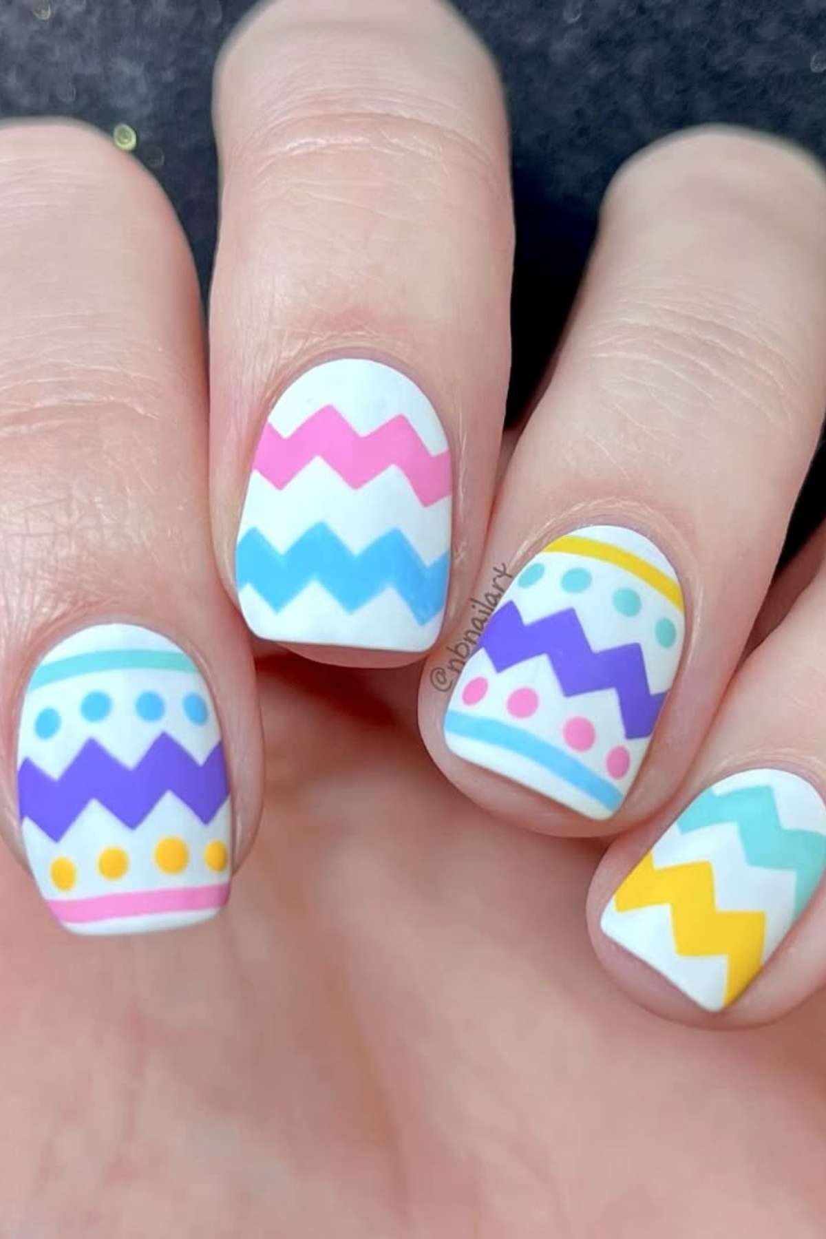 A hand with fingernails painted white and decorated with colorful zigzag and dot patterns in pastel shades of blue, pink, yellow, and purple.