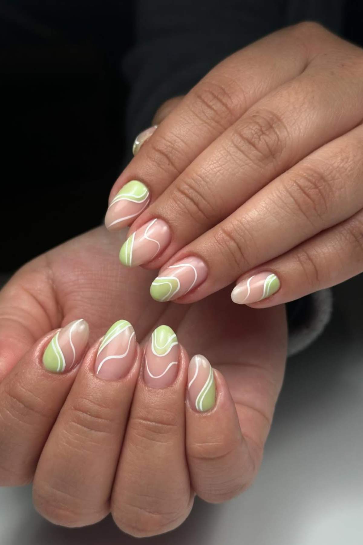 Hands with almond-shaped nails featuring a nude base and abstract green and white wavy line designs.