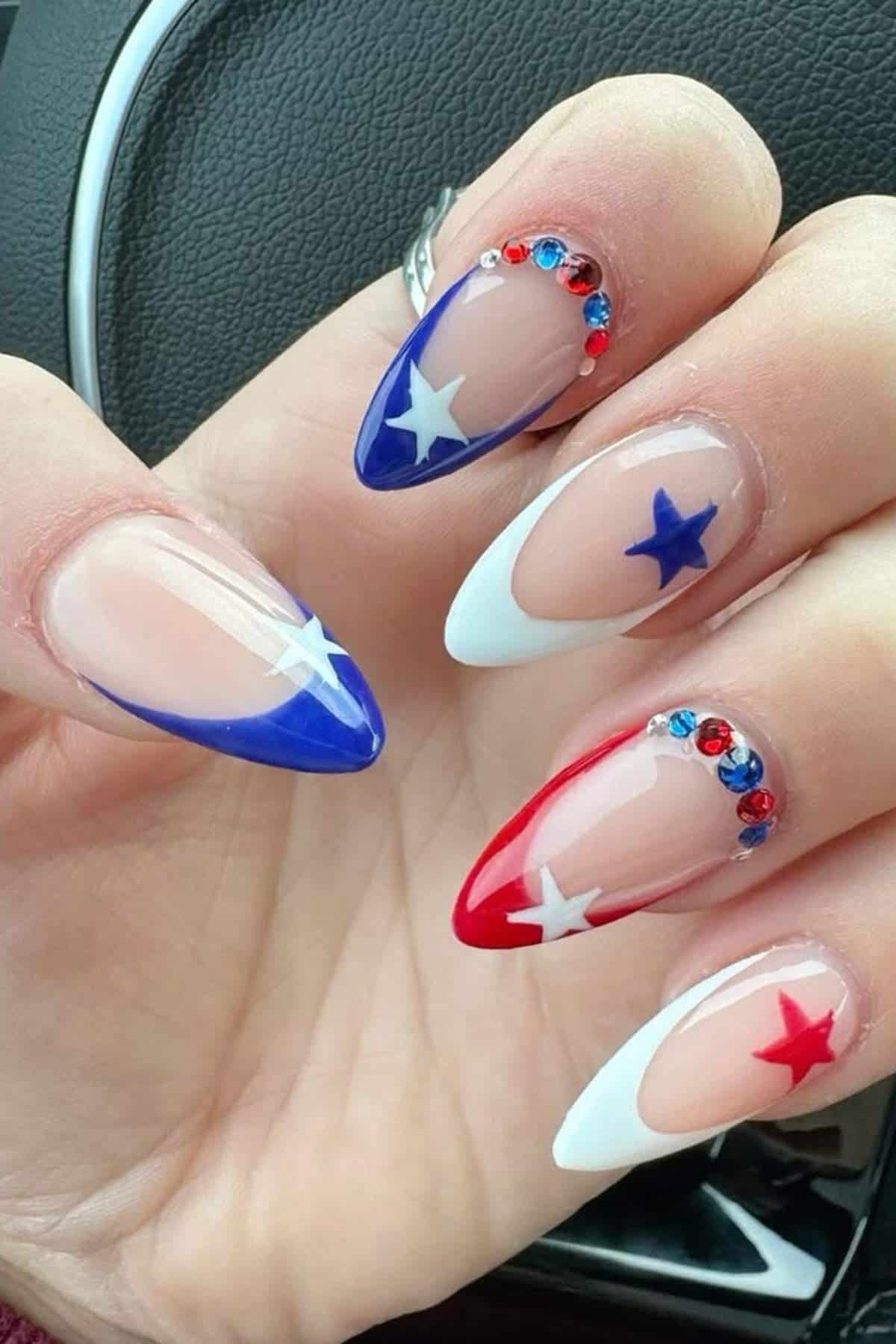 A hand with long, pointed nails painted in red, white, and blue with star designs and small rhinestones, against a car interior background.