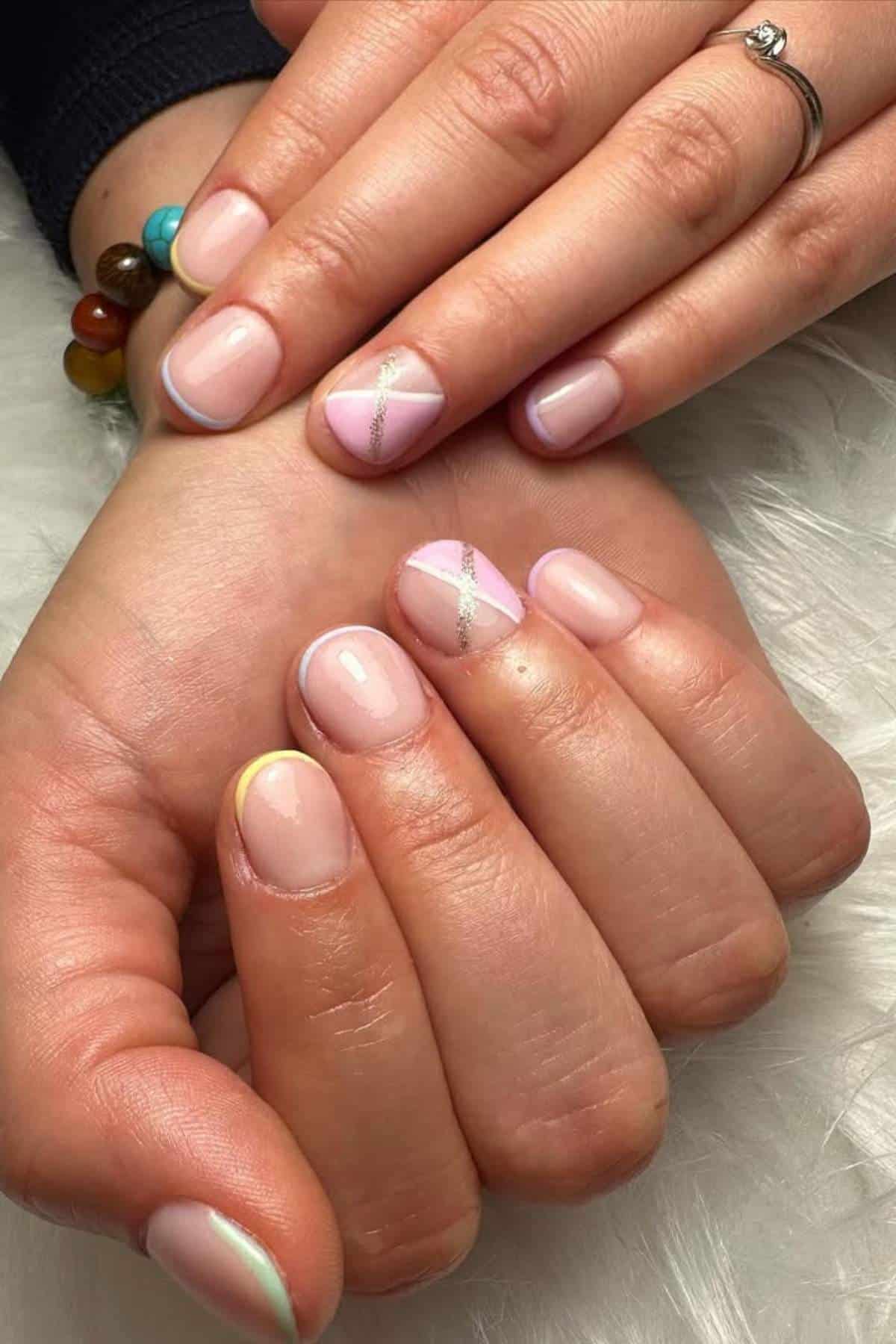 Hands with short, natural-shaped nails painted in a pale pink polish. Some nails feature pastel diagonal lines and shimmer accents. A ring and colorful bracelet are visible on the wrist.