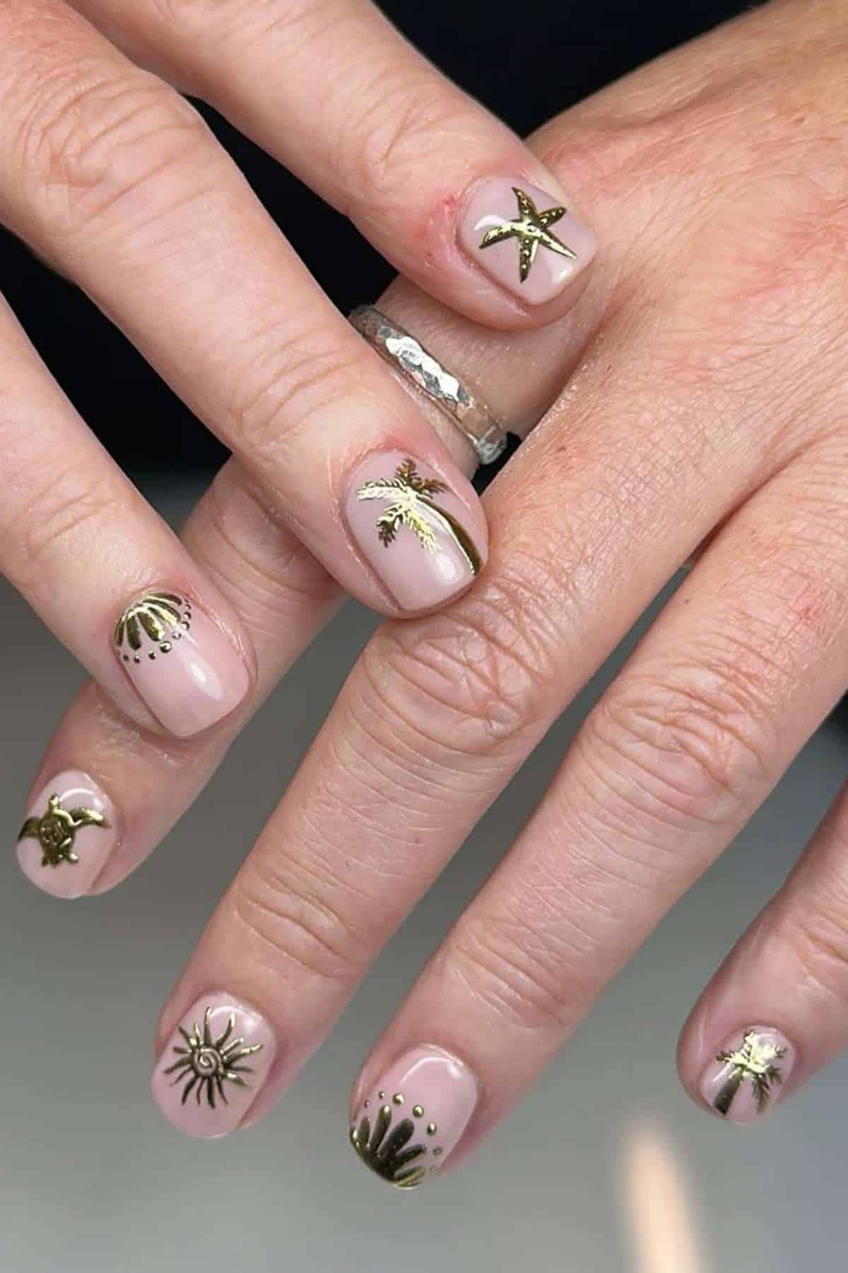 Hands with short, natural nails painted nude, each nail decorated with different small gold designs, including a starfish, shell, palm tree, sun, and abstract patterns.