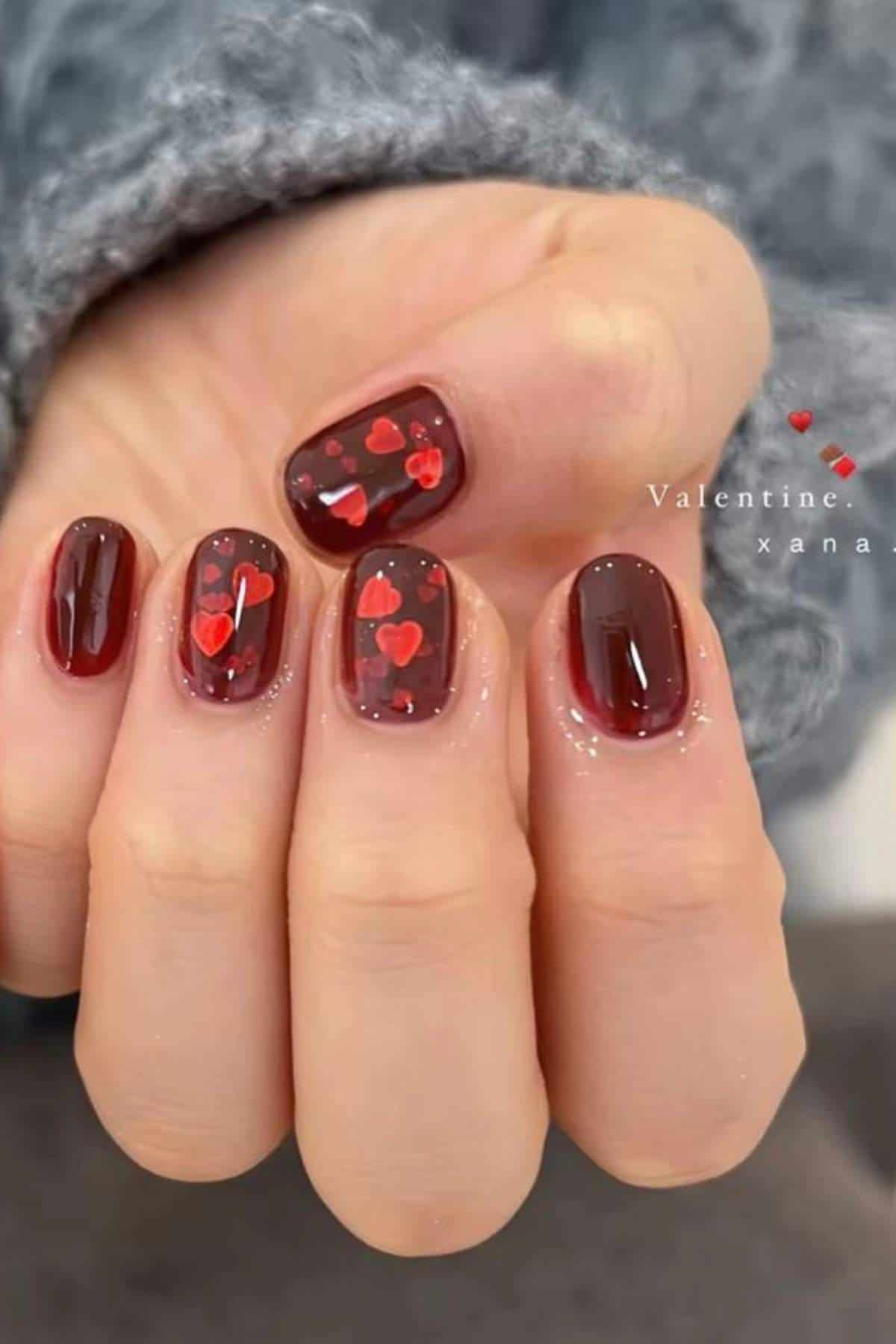 A hand with short, dark red nails featuring small red and pink heart designs on some fingers.