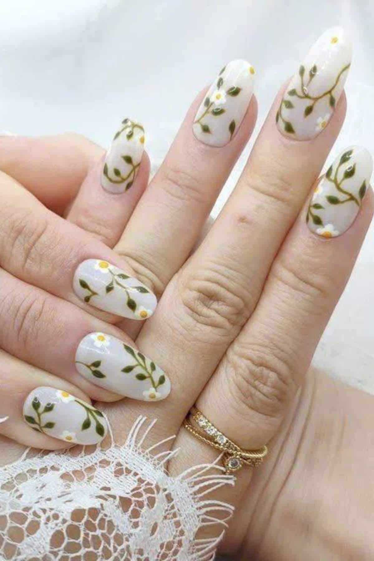Hands with long, almond-shaped nails painted with white polish and green vine and small white flower designs; person wears two gold rings.