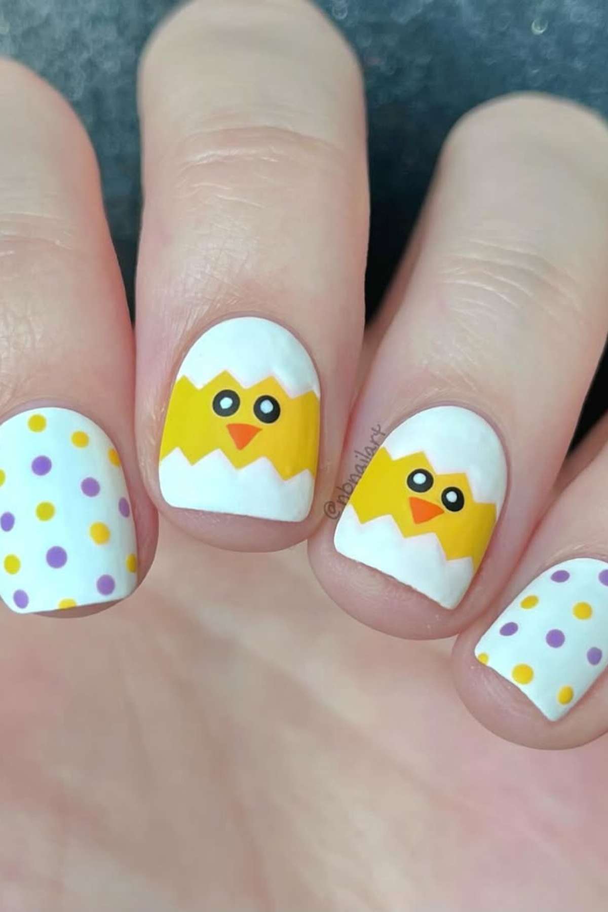 Fingernails painted with white polish, featuring yellow chick faces on two nails and purple and yellow polka dots on the others.