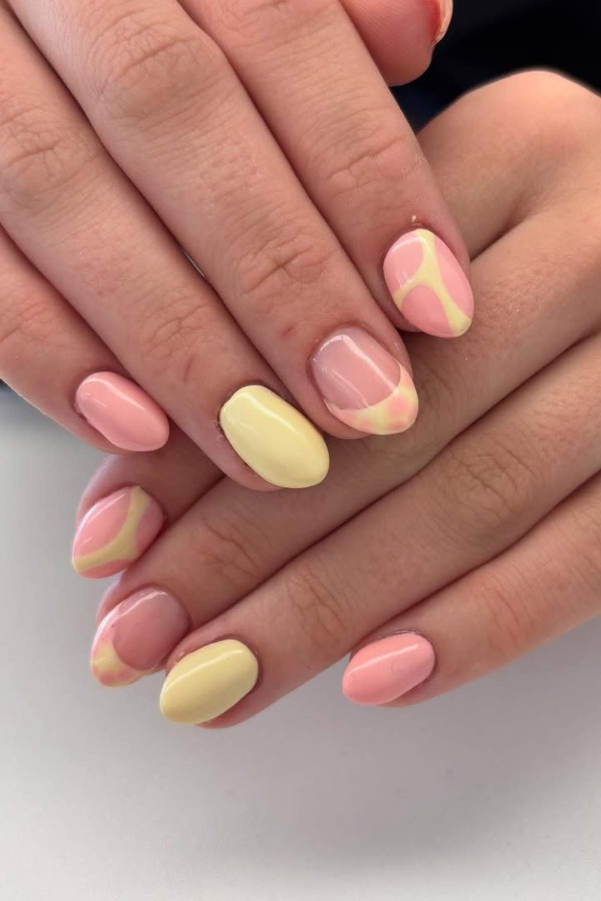 Hands with almond-shaped nails painted in pastel yellow and pink, featuring abstract swirl designs on some nails.