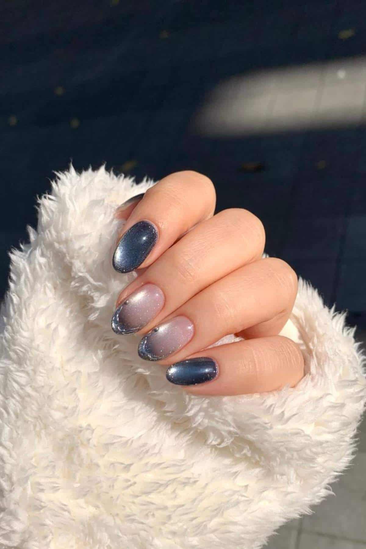 A hand with short, oval nails features a gradient manicure in blue and nude shades with a subtle shimmer, shown against a white, fuzzy sleeve.