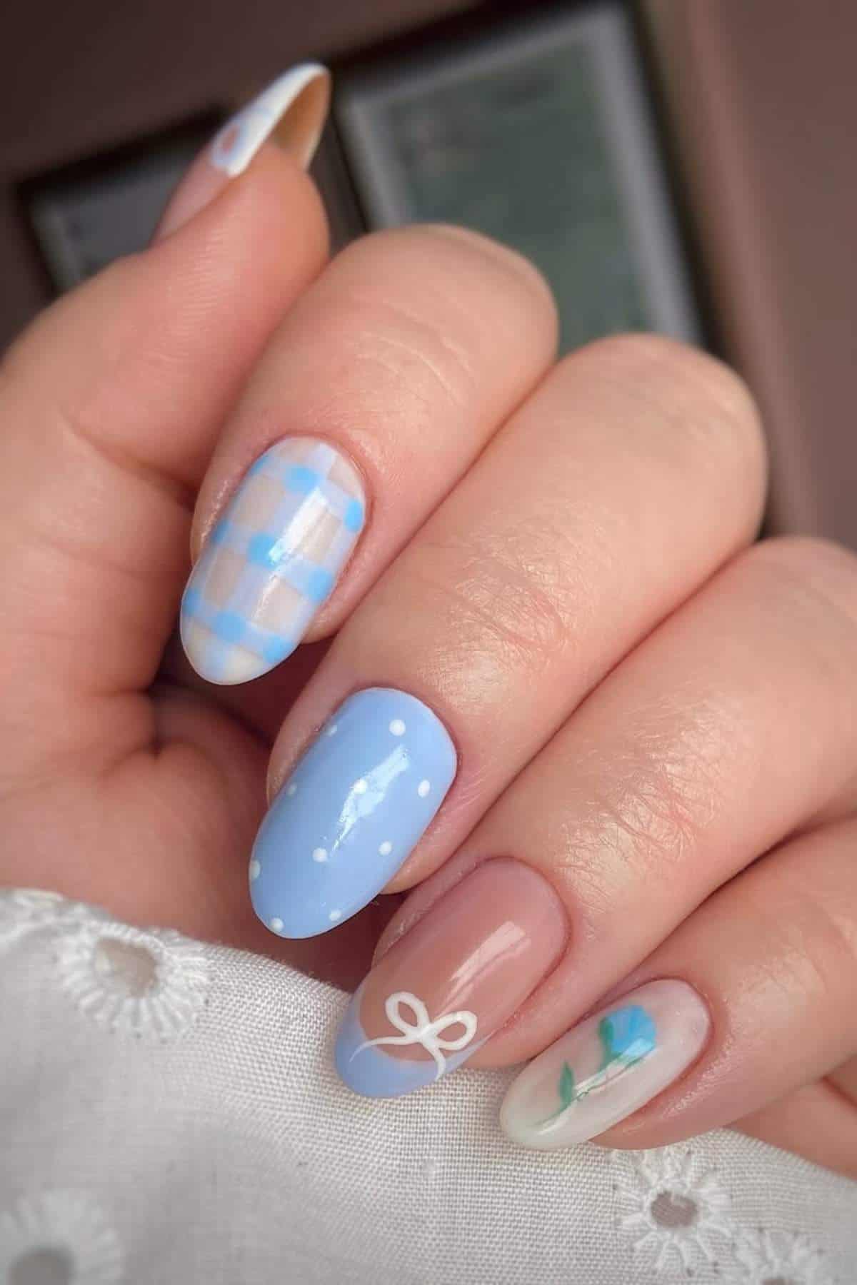 A hand with manicured nails featuring different designs: blue plaid, solid blue with white dots, nude with a white bow, and nude with blue and green floral accents.
